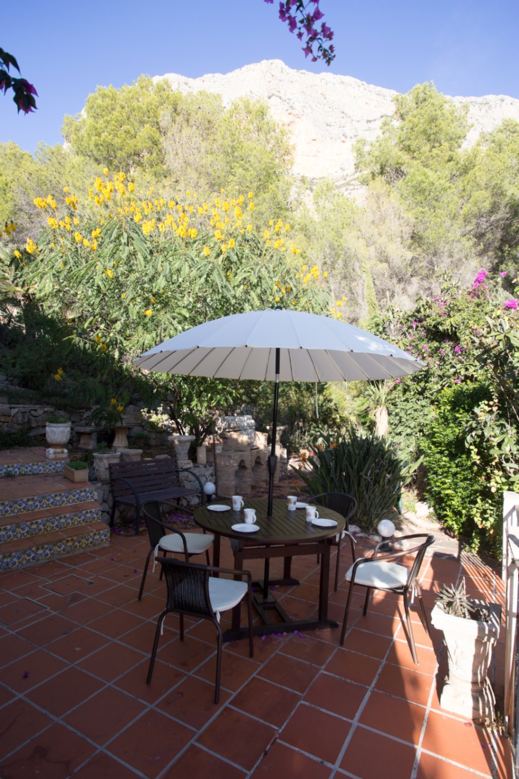 Terracotta terras met een ronde houten eethoek en witte parasol, omgeven door een mediterrane tuin met uitzicht op een rotsachtige bergwand.