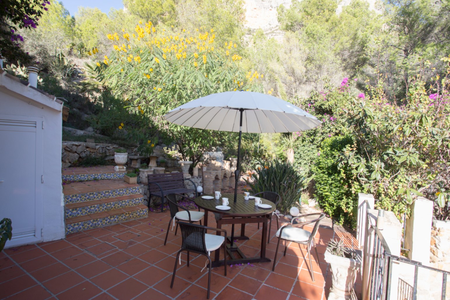 Terras met terracotta tegels, een houten eethoek onder een witte parasol en decoratieve betegelde trappen tegen een beboste heuvelwand.