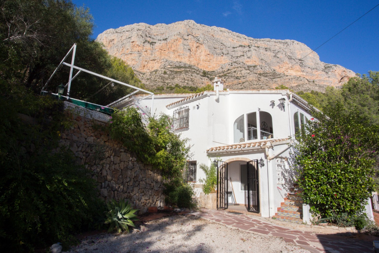 Witte mediterrane villa met boogvensters aan de voet van een rotsachtige berg, voorzien van natuurstenen muren en een betegelde buitentrap.