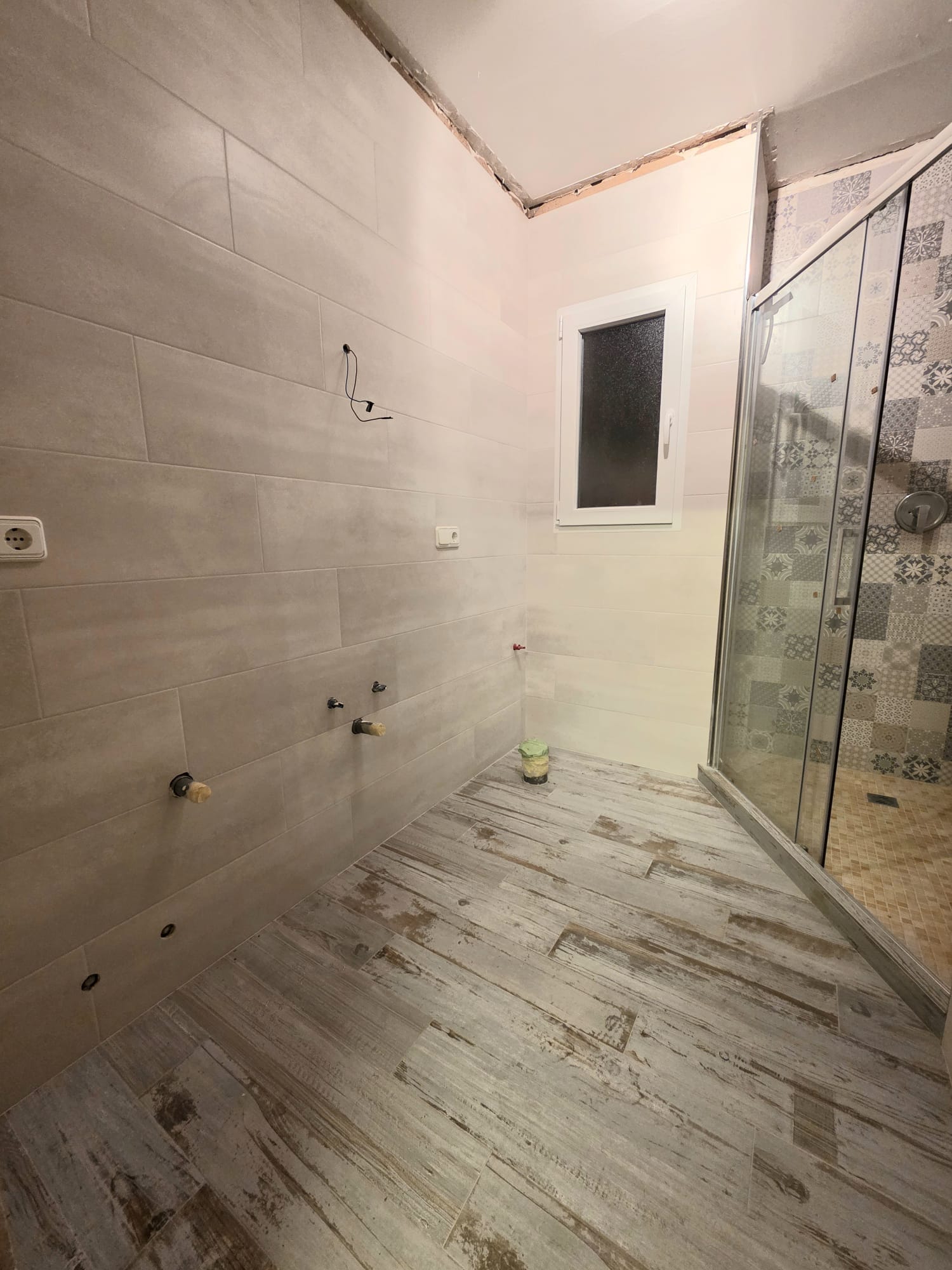 Badkamer in renovatie met houtlook vloertegels, grootformaat wandtegels en een glazen douchewand voorzien van decoratieve patroontegels.