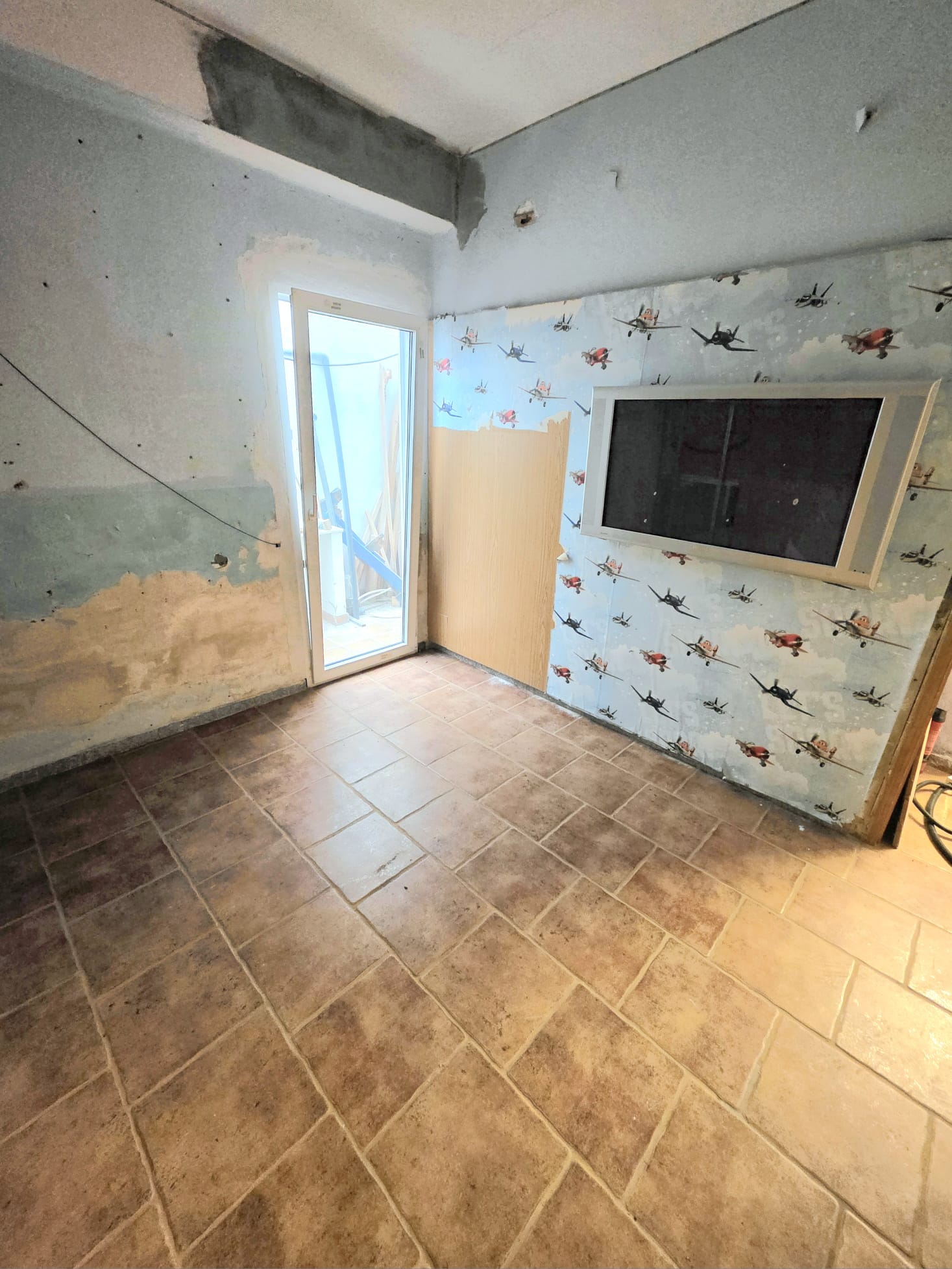 Binnenruimte in renovatie met getextureerde vloertegels, een witte glazen deur en een wand met vliegtuigbehang en een ingebouwd frame voor een beeldscherm.