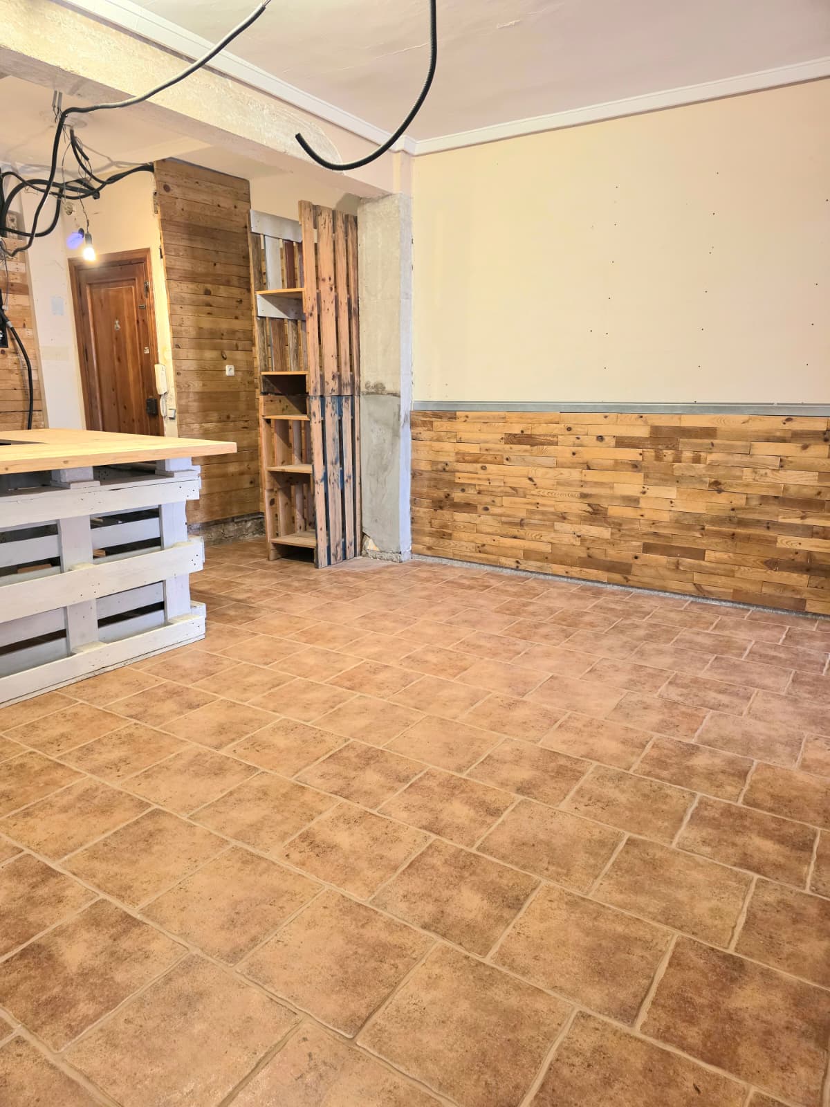 Binnenruimte met terracotta tegelvloer en rustieke houten wandpanelen. Voorzien van een witte palletbalie en houten stellingkasten voor een industrieel accent.