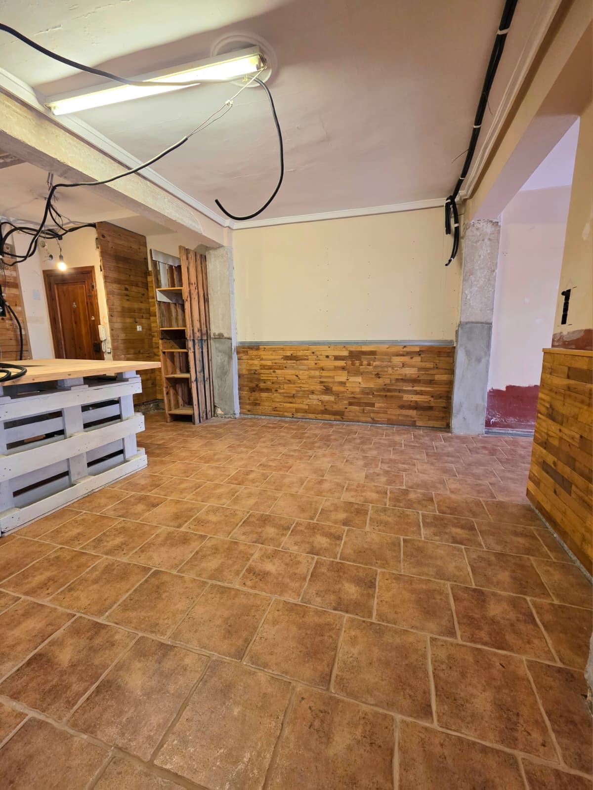 Binnenruimte met terracotta tegelvloer, rustieke houten wandpanelen en een balie gemaakt van witgeschilderde pallets met een licht houten werkblad.