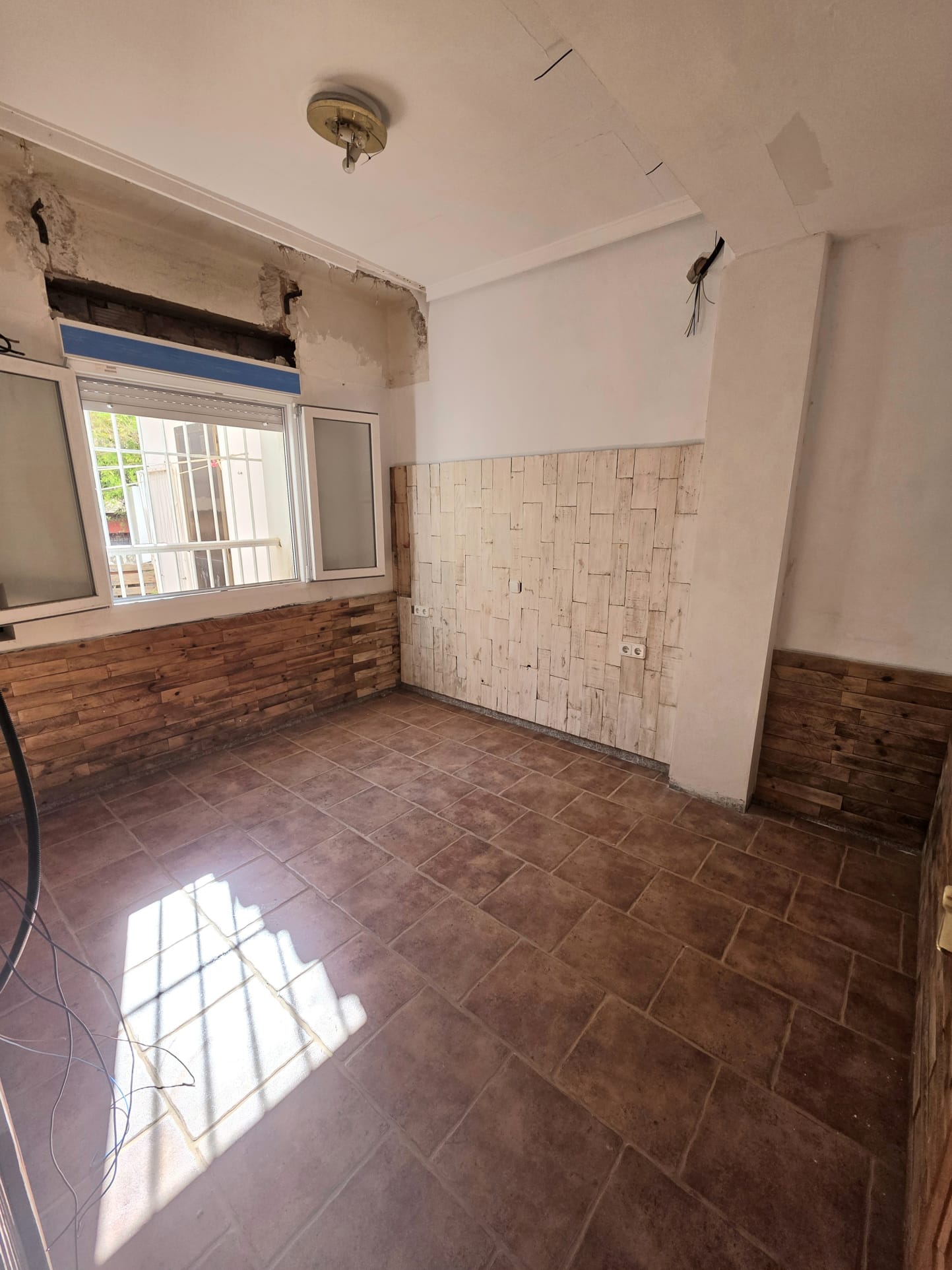 Kamer met terracotta tegelvloer en decoratieve houten wandbekleding. Natuurlijk licht valt binnen via het witte raamkozijn in deze ruimte in renovatie.