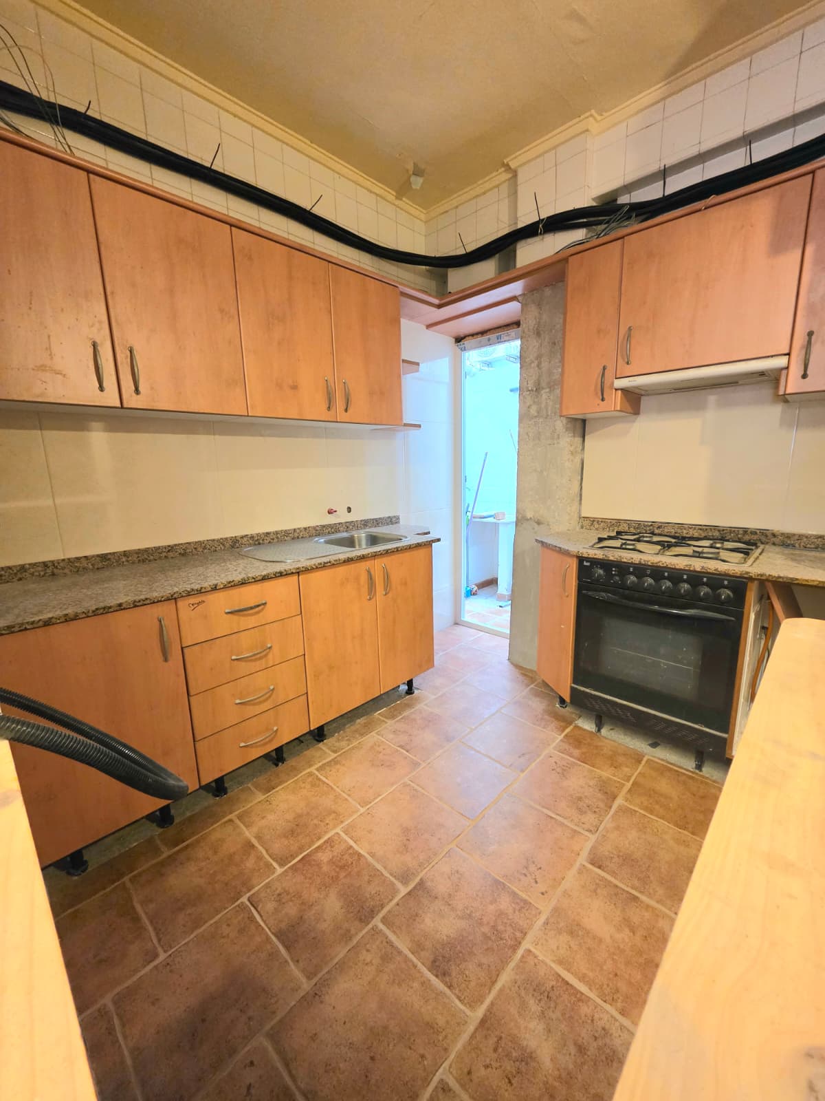 Keuken met houten kasten, granieten aanrechtblad en terracotta tegelvloer. Voorzien van een zwarte oven, gaskookplaat en toegang tot een aangrenzende wasruimte.