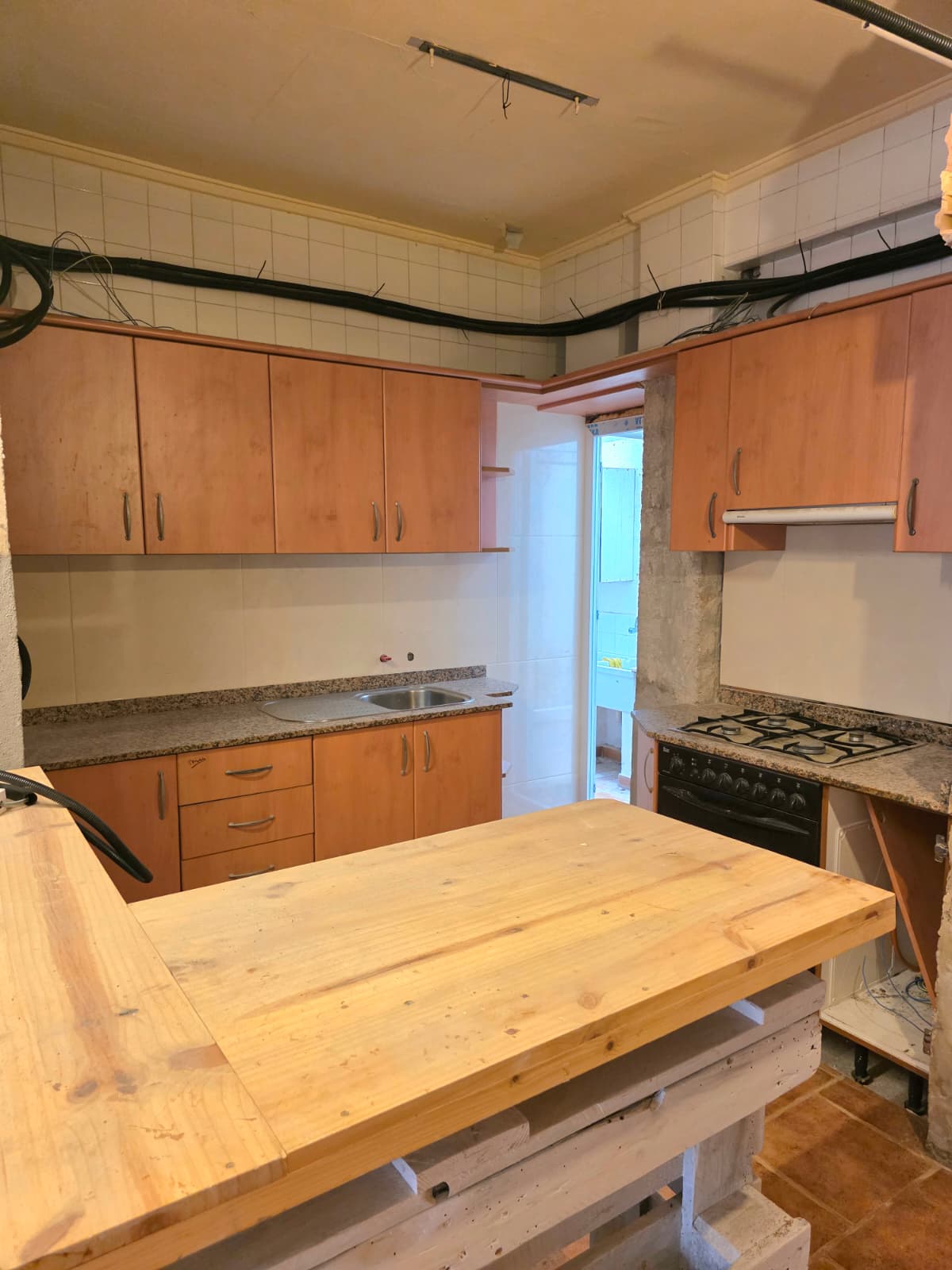 Keuken met houten kasten, granieten werkbladen en een centraal houten kookeiland. Voorzien van betegelde wanden en zichtbare leidingen aan de muur.