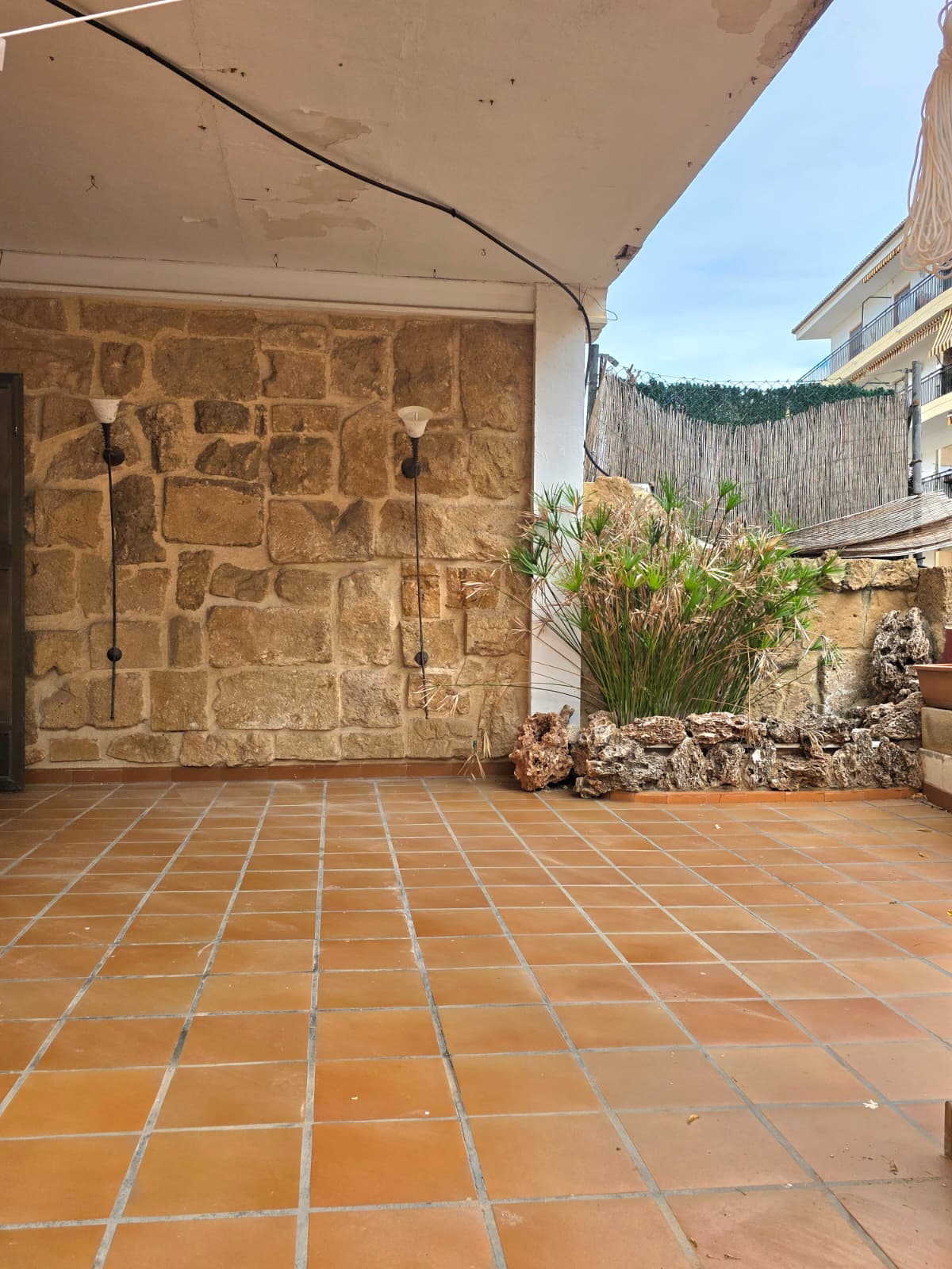 Overdekt terras met terracotta tegelvloer, een rustieke natuurstenen muur voorzien van wandverlichting en een decoratieve plantenbak met rotsrand.