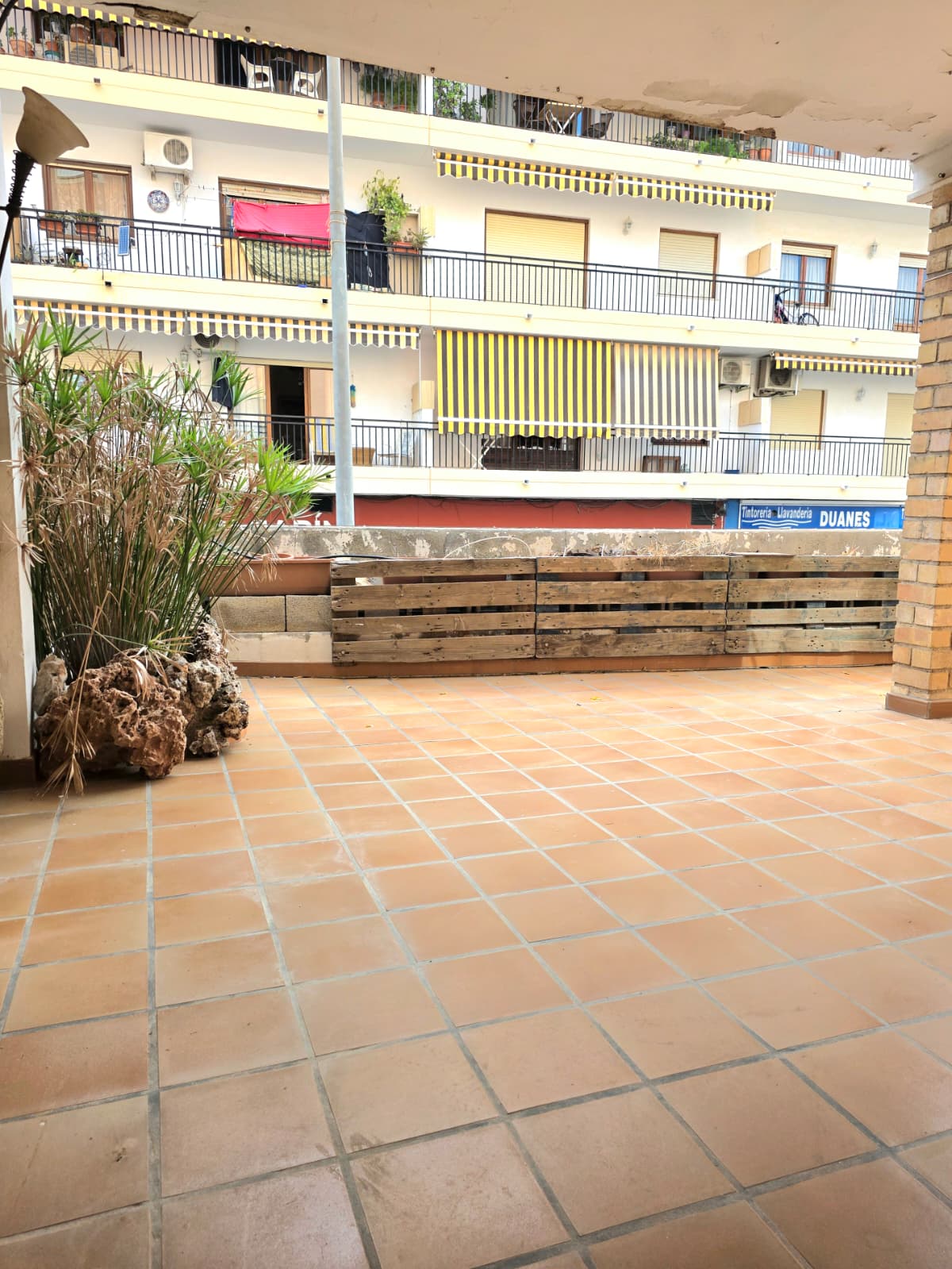 Ruim buitenterras met terracotta tegelvloer, een afscheiding van houten pallets en uitzicht op omliggende gebouwen met gestreepte zonneschermen.