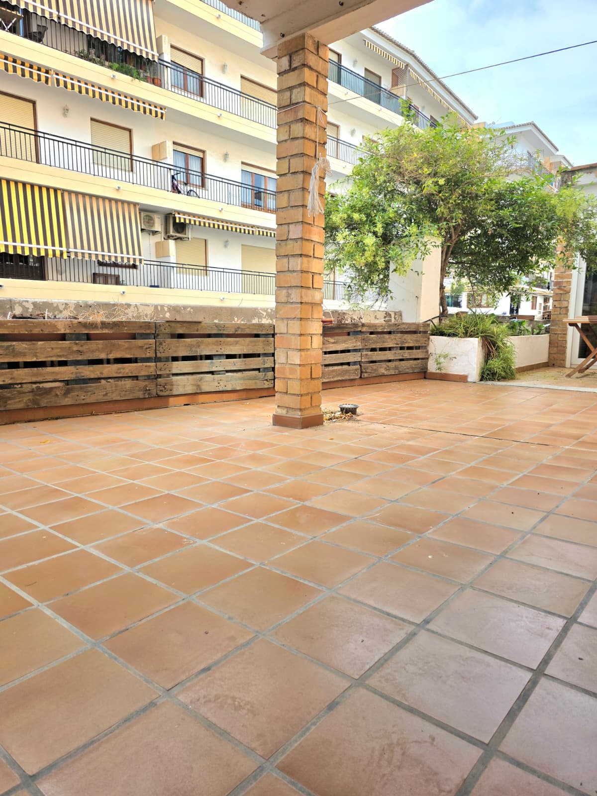 Ruim buitenterras met terracotta tegelvloer, een centrale bakstenen pilaar en houten palletafscheidingen met geïntegreerd groen.