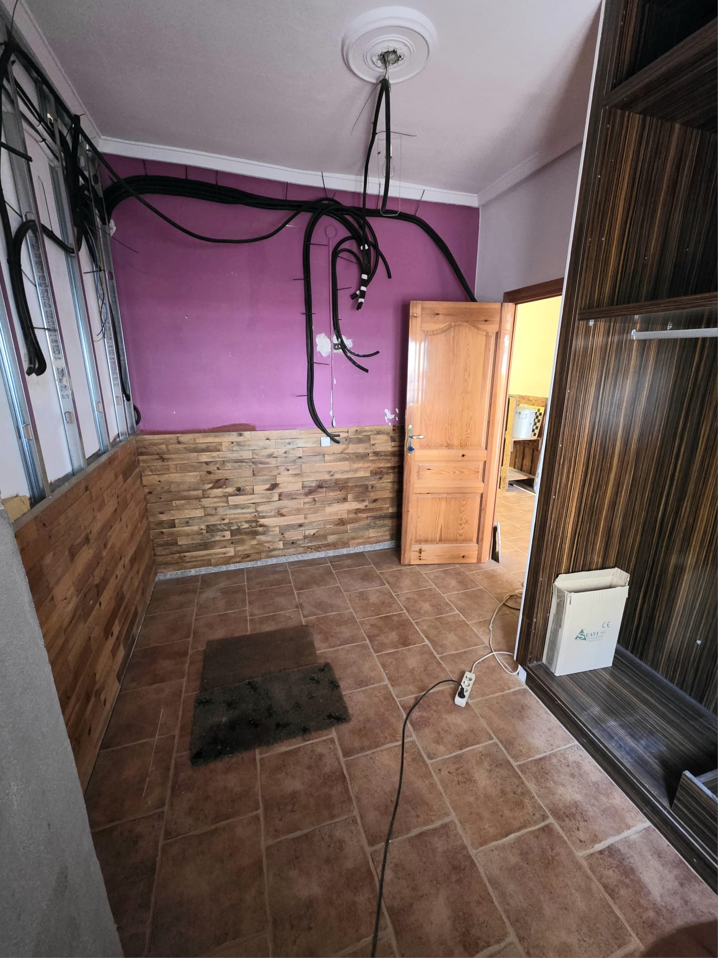 Te renoveren kamer met terracotta vloertegels, houten wandlambrisering, paarse muren en zichtbare elektrische bedrading en metalen profielen.