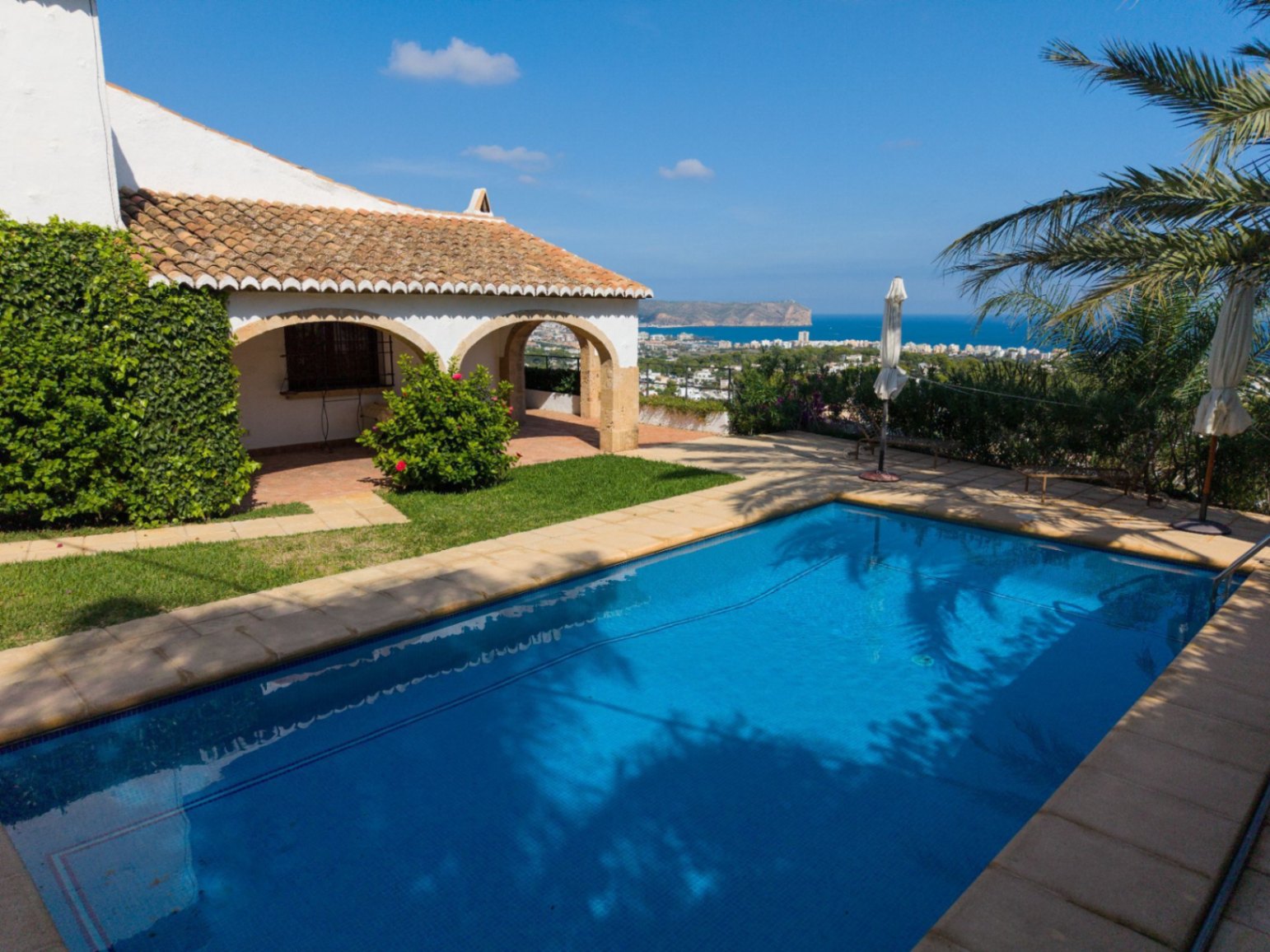 Buitenzwembad met stenen rand naast een mediterrane villa met een overdekt terras met bogen en een pannendak, met uitzicht op de zee en de kustlijn.