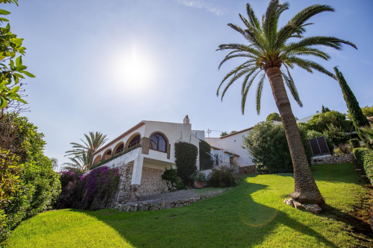 Exterieur van een mediterrane villa met een overdekt boogterras, natuurstenen muren en een glooiend gazon met een karakteristieke palmboom.