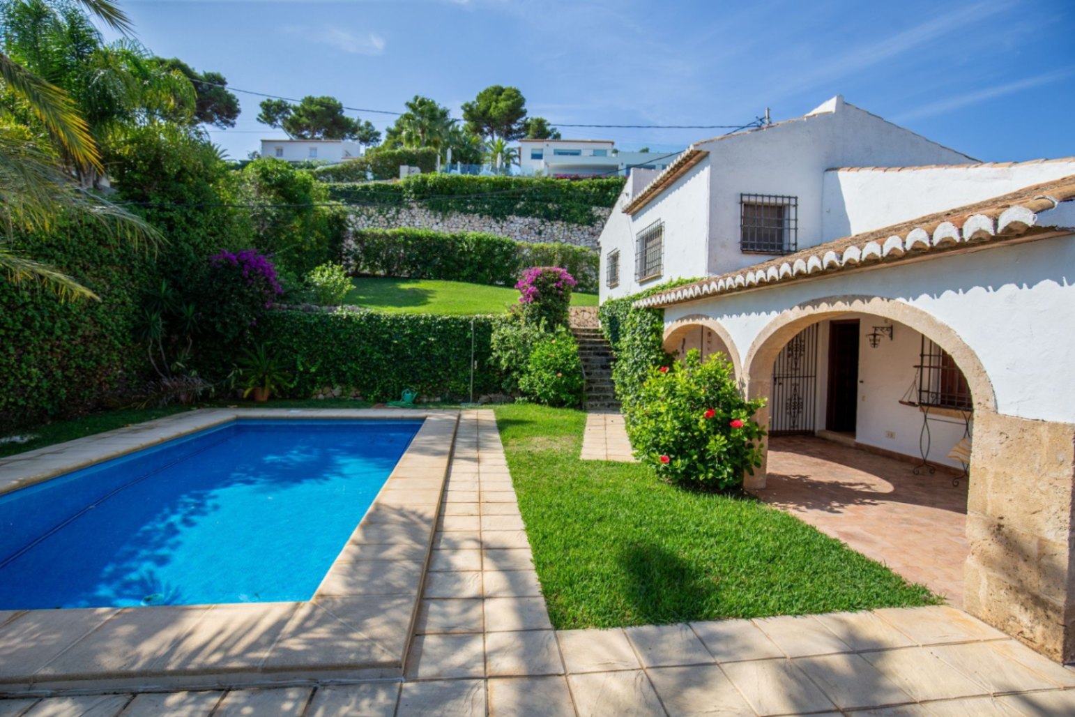 Exterieur van een mediterrane villa met rechthoekig zwembad, stenen boogportiek en gelaagde tuinen met volwassen beplanting en bougainville.