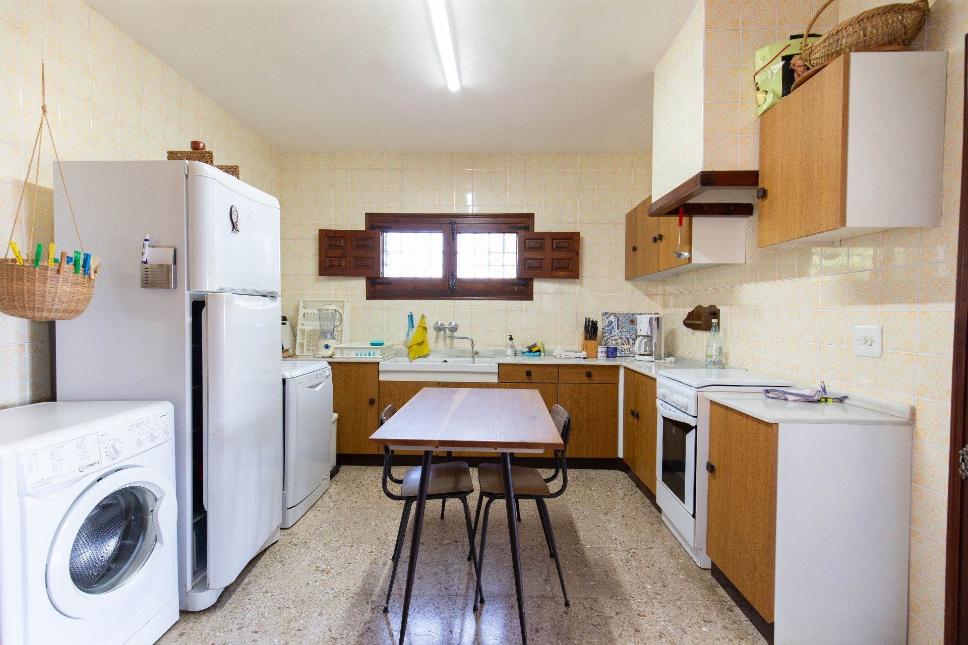 Functionele keuken met houten kasten, gedessineerde wandtegels en witgoed. Een centrale houten eettafel staat op een terrazzovloer bij een raam met houten luiken.