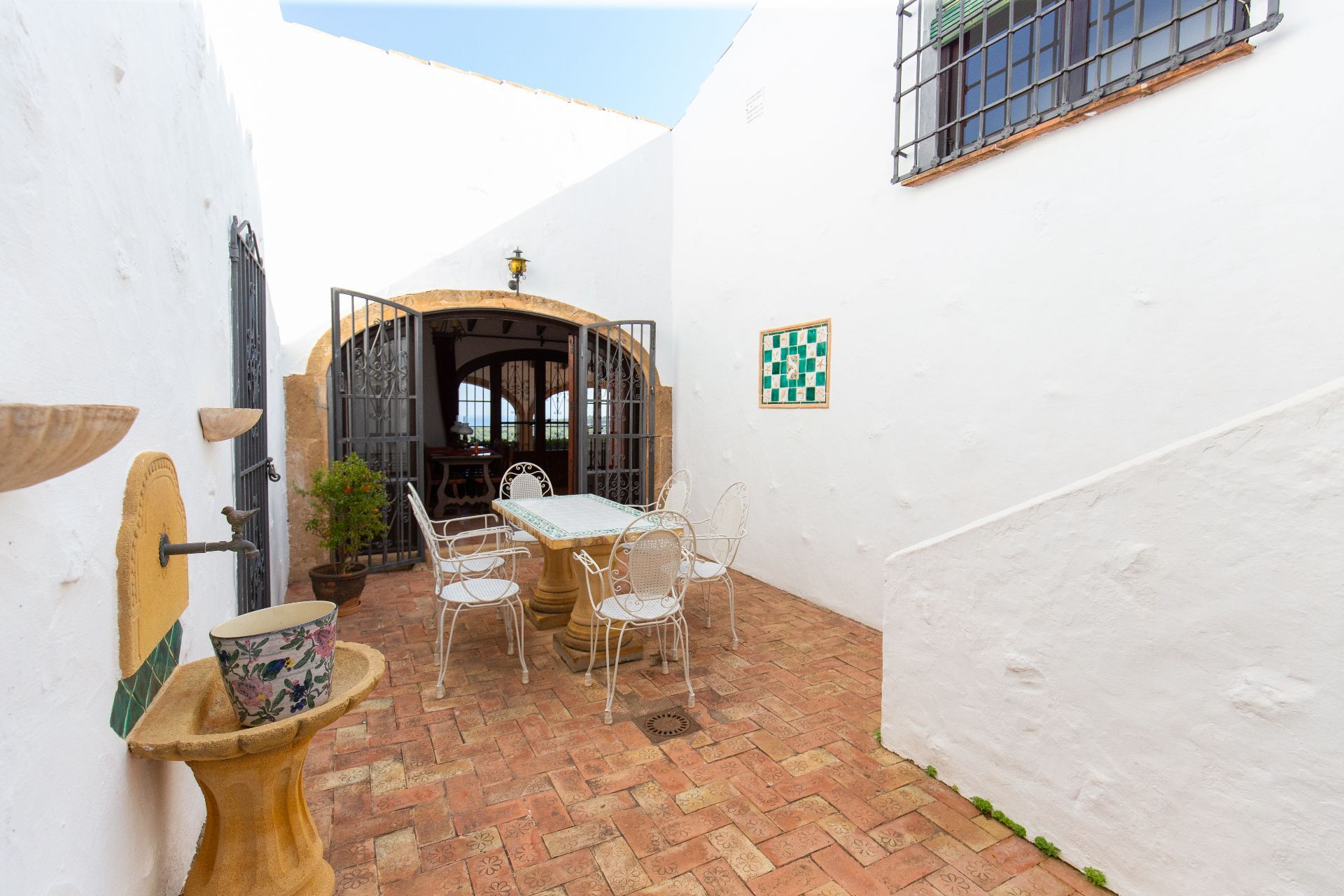 Mediterrane patio met gedecoreerde terracotta tegels, een stenen fontein en wit smeedijzeren meubilair. Een boogingang met sierhekwerk leidt naar het interieur.