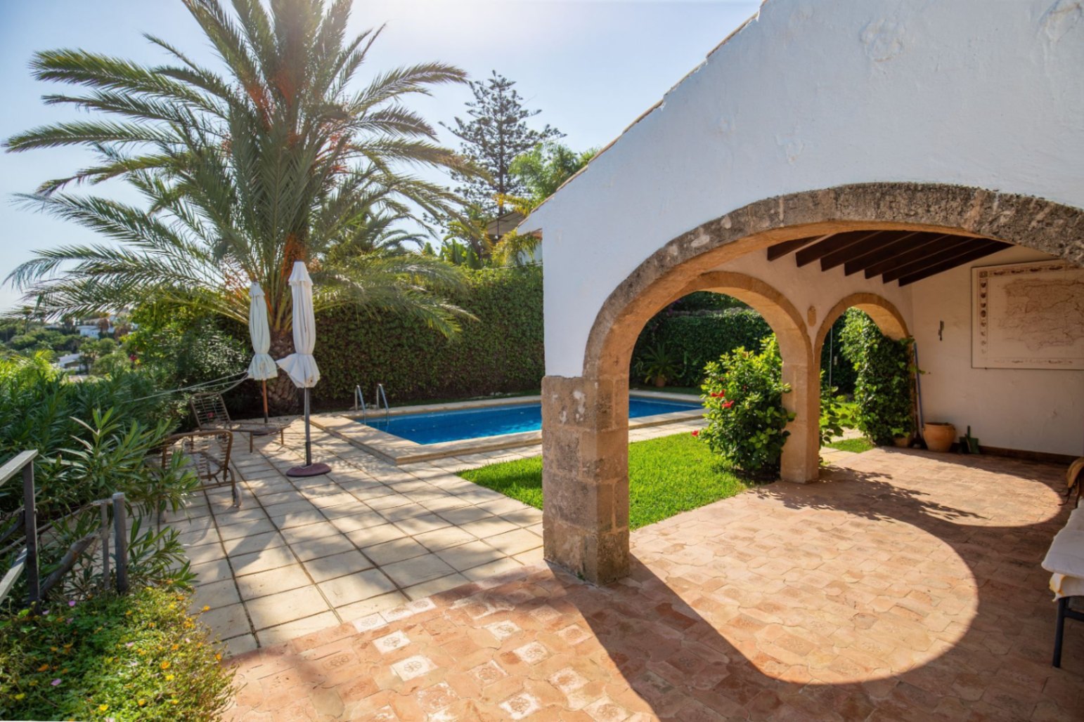 Overdekt terras met stenen bogen en terracotta vloer met uitzicht op een privézwembad, omgeven door palmbomen en een volgroeide mediterrane tuin.