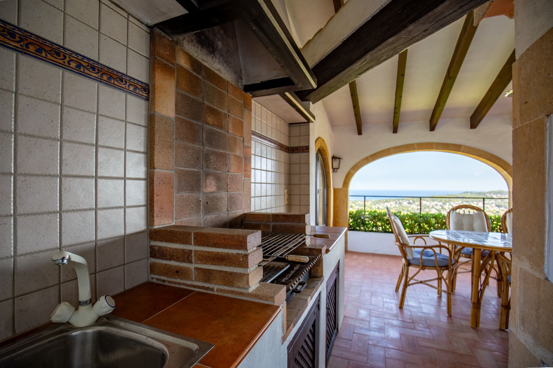 Overdekte buitenkeuken met een gemetselde barbecue, gootsteen en terracotta vloer, met houten balkenplafonds en uitzicht op zee door een boog.