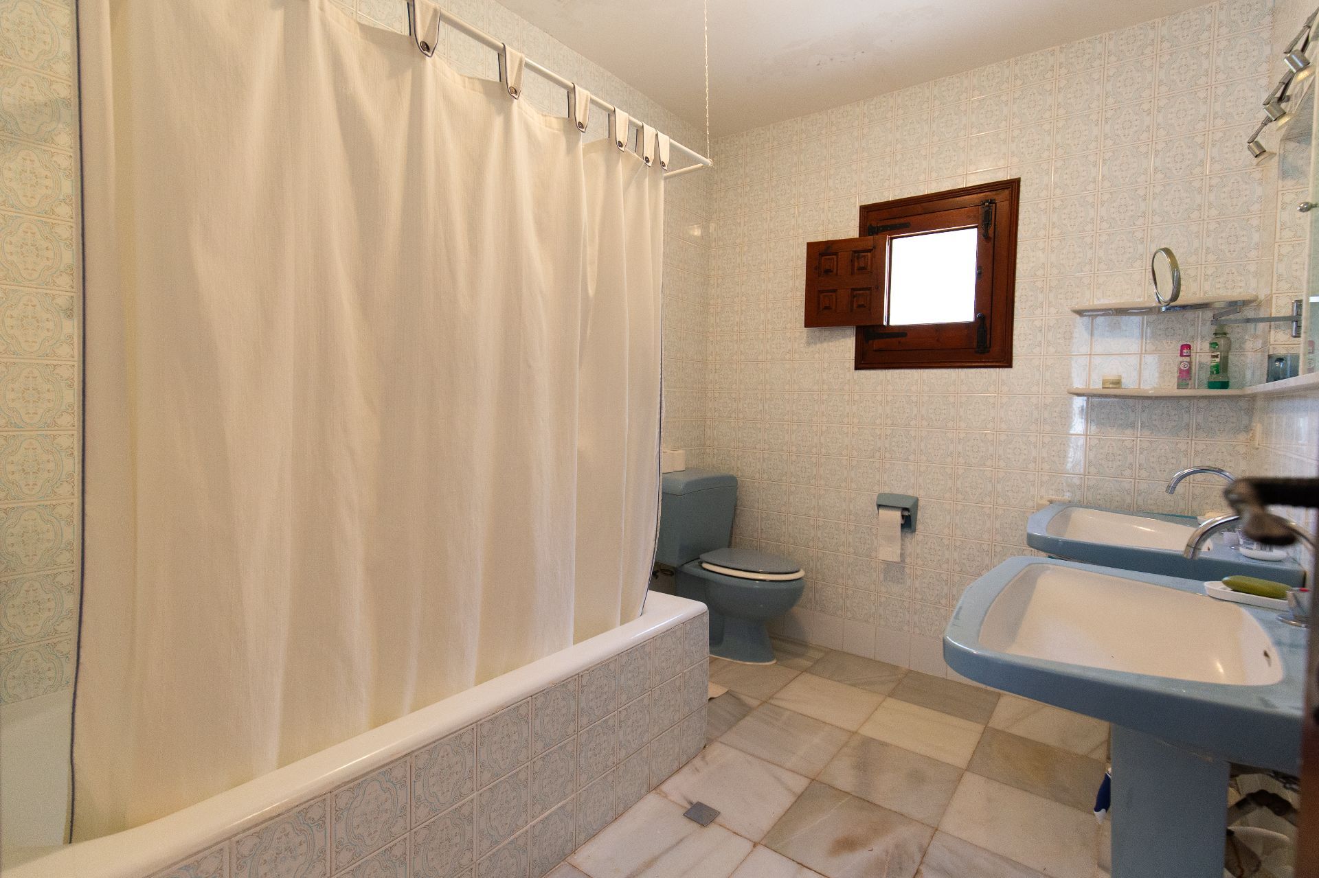 Traditionele badkamer met dubbele blauwe wastafels op zuil, bijpassend toilet en een betegeld ligbad. Voorzien van gedecoreerde wandtegels en een houten venster.