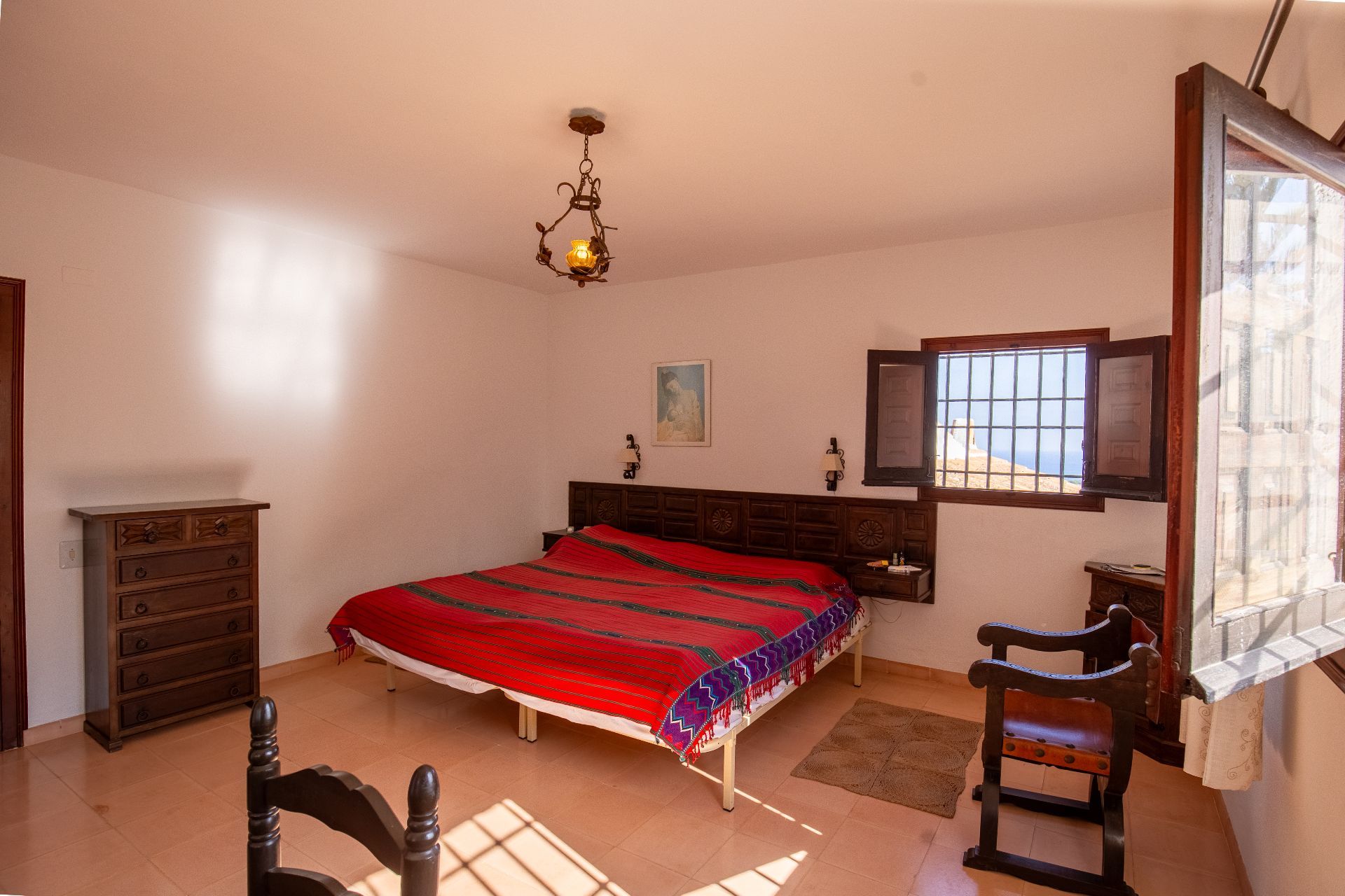 Traditionele slaapkamer met een bewerkt houten hoofdbord, terracotta vloertegels en een smeedijzeren kroonluchter. Een venster met houten luiken biedt natuurlijk lichtinval.