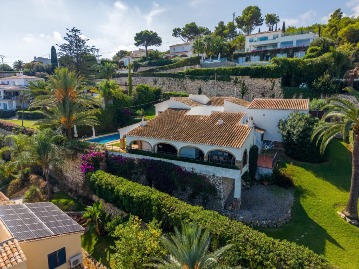 Verhoogd zicht op een mediterrane villa met terracotta daken, boogterras en privézwembad, omgeven door stenen muren en tuinen met palmbomen.