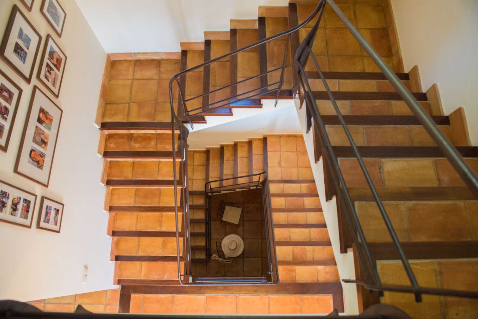 Bovenaanzicht van een trappenhuis met terracotta treden, donkerhouten afwerking en een smeedijzeren leuning. De witte wand is gedecoreerd met diverse ingelijste foto's.