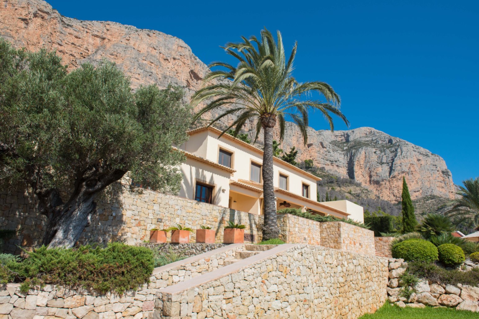 Exterieur van een mediterrane villa met natuurstenen muren, een olijfboom en een palmboom tegen een achtergrond van indrukwekkende rotswanden.