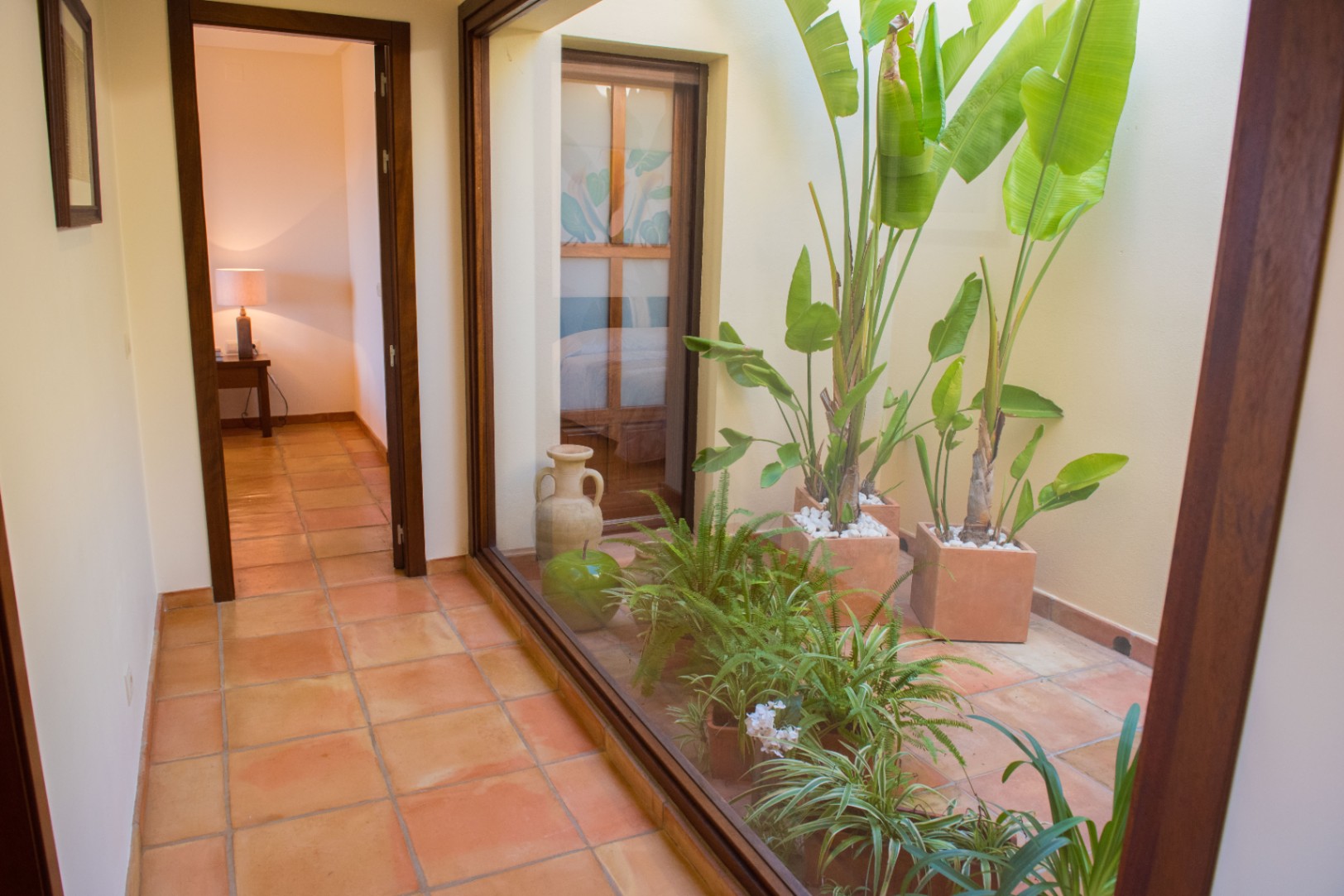 Gang met terracotta vloertegels en een grote glazen wand die uitkijkt op een interne patio met tropische planten.