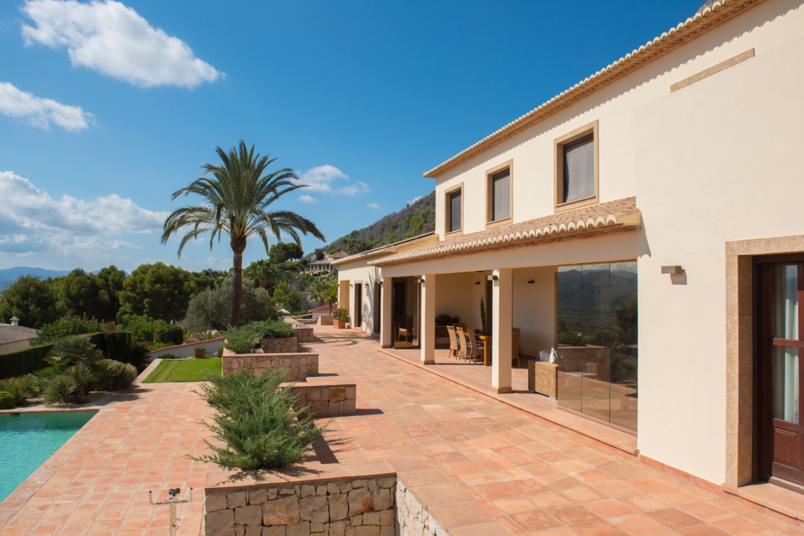 Mediterrane villa met een terracotta terras, natuurstenen borders en een overdekte veranda. Een palmboom siert de buitenruimte bij het zwembad en de bergen.