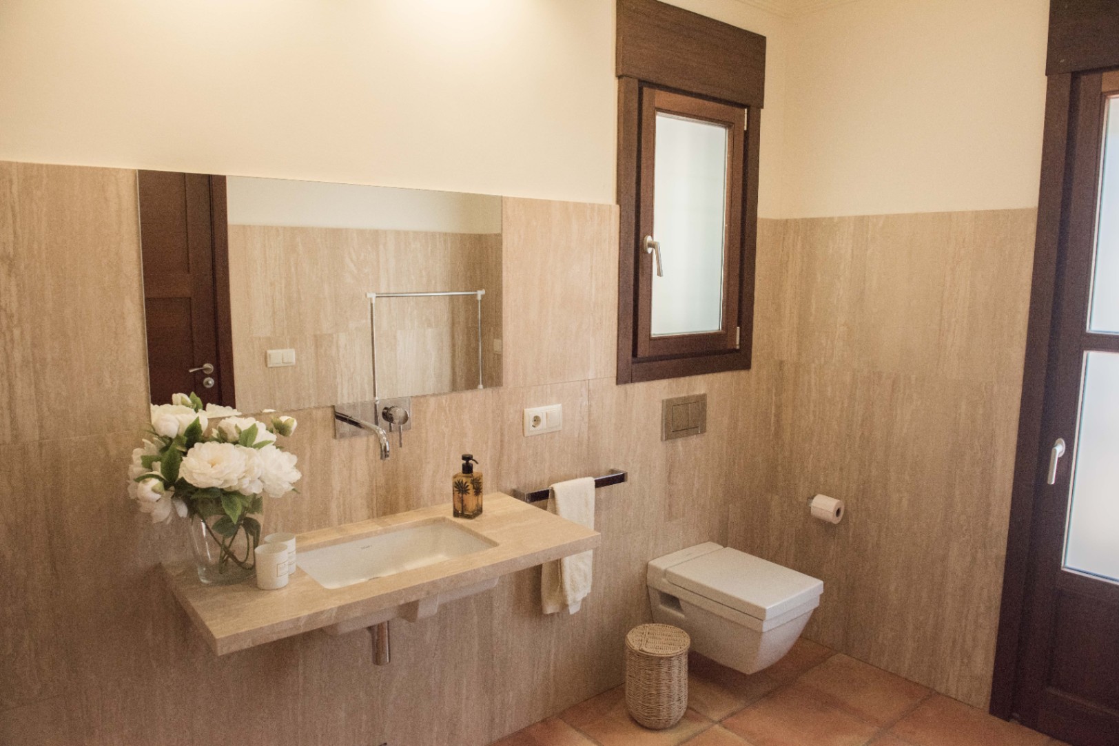 Moderne badkamer met een zwevend natuurstenen wastafelblad, inbouwkranen en een hangend toilet tegen wanden van travertijn en een terracotta vloer.