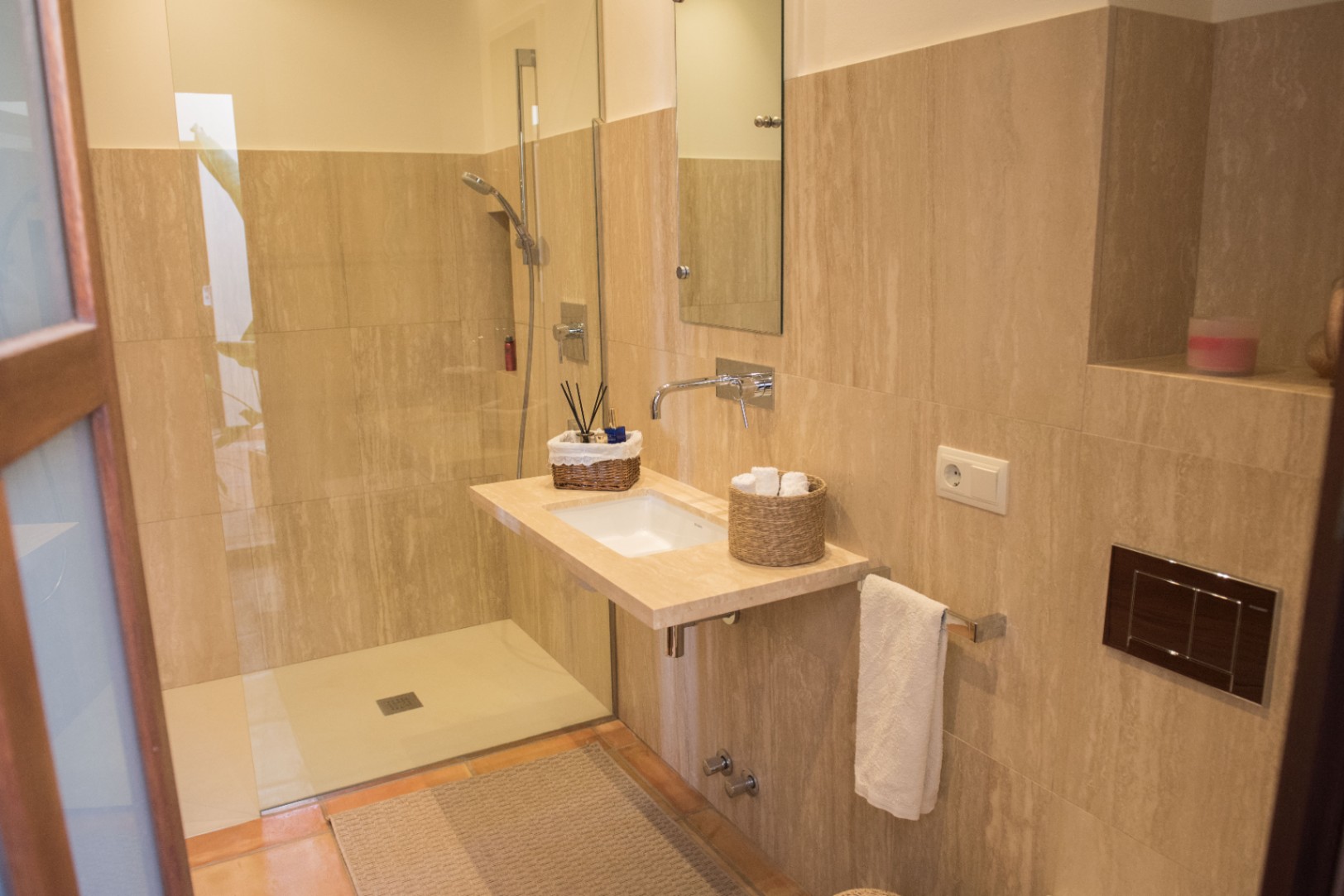 Moderne badkamer met inloopdouche, zwevend natuurstenen wastafelblad en inbouwkranen tegen een achtergrond van beige travertijn tegels.