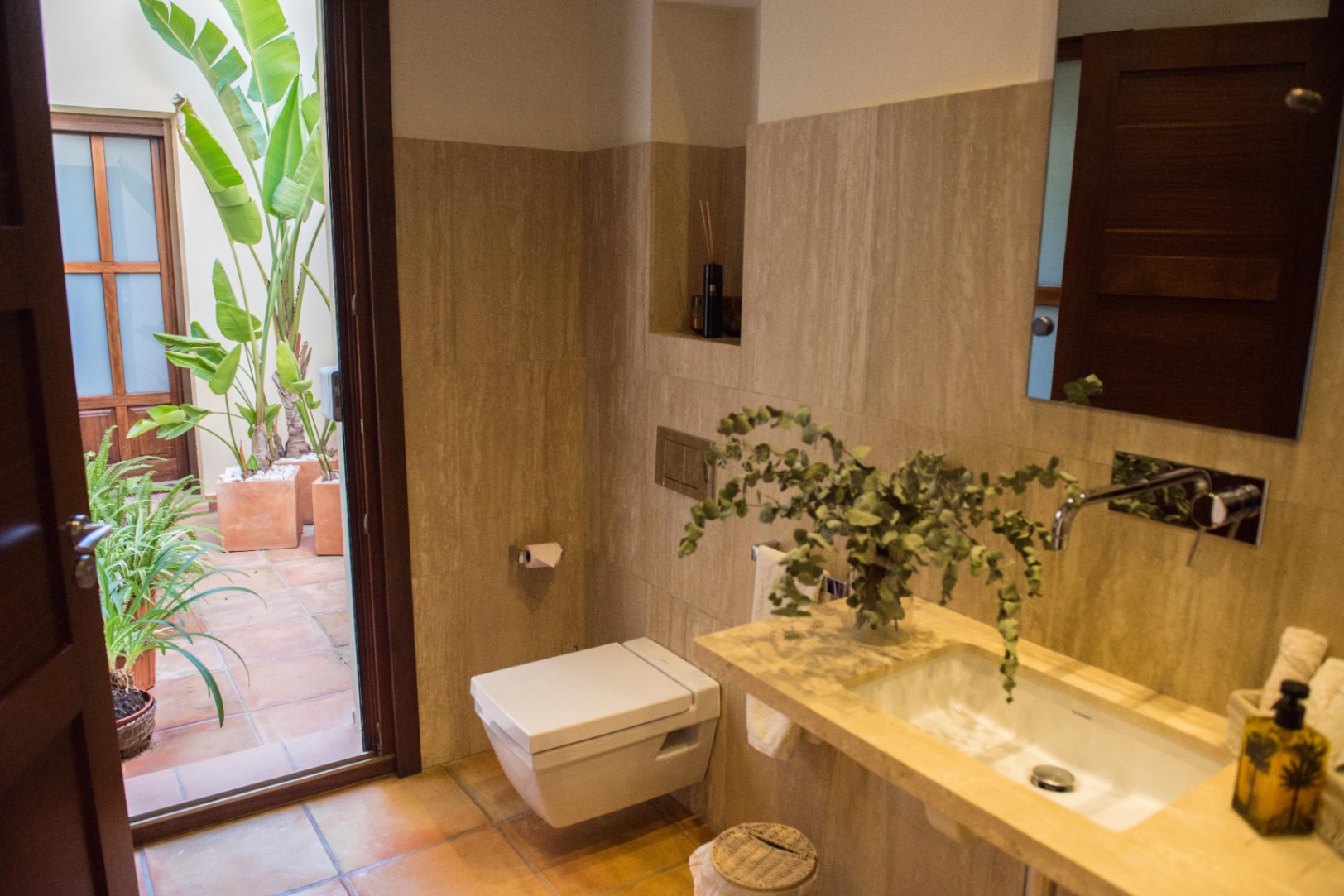 Moderne badkamer met travertijn wandafwerking, hangend toilet en directe toegang tot een privépatioterras met terracotta tegels en tropische planten.