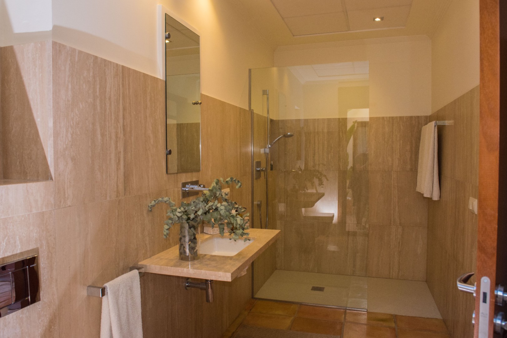 Moderne badkamer met travertijn wandtegels, een zwevend stenen wastafelmeubel en een inloopdouche met glazen wand en terracotta vloer.