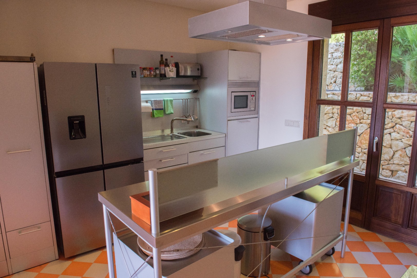Moderne keuken met rvs-eiland, geblokte vloertegels en houten kozijnen die uitkijken op een karakteristieke natuurstenen buitenmuur.
