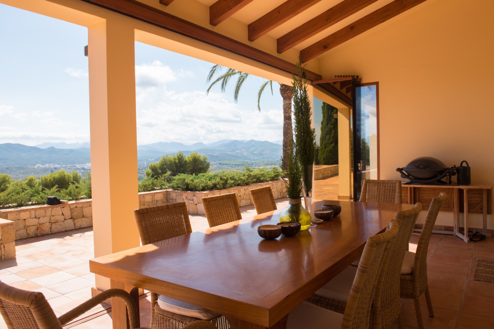 Overdekt terras met een grote houten eettafel en rotan stoelen onder een balkenplafond. Panoramisch uitzicht op de bergen en een terras met terracotta tegels.