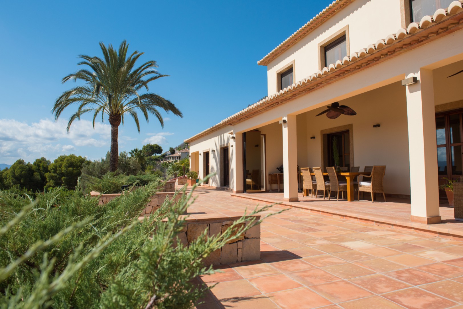 Terracotta terras met een overdekte eethoek en rieten stoelen, uitkijkend op een palmboom en de omliggende mediterrane beplanting.