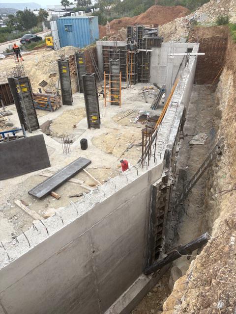 Funderingsfase met steunmuren van gewapend beton en verticale bekistingen voor structurele kolommen op een uitgegraven bouwterrein.