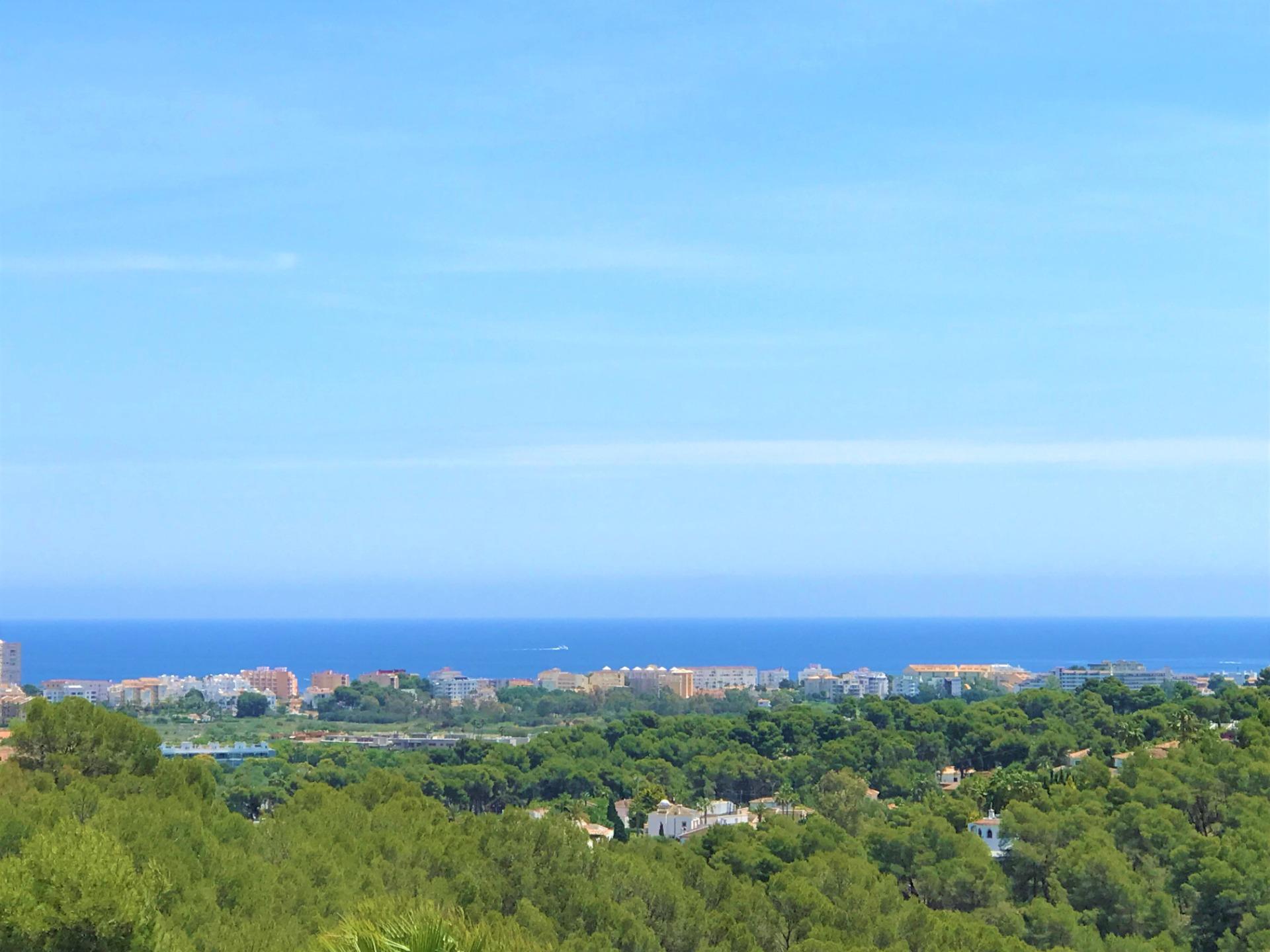 Panoramisch uitzicht op de Middellandse Zeekust, met een dicht dennenbos op de voorgrond en kustarchitectuur tegen een helderblauwe horizon.