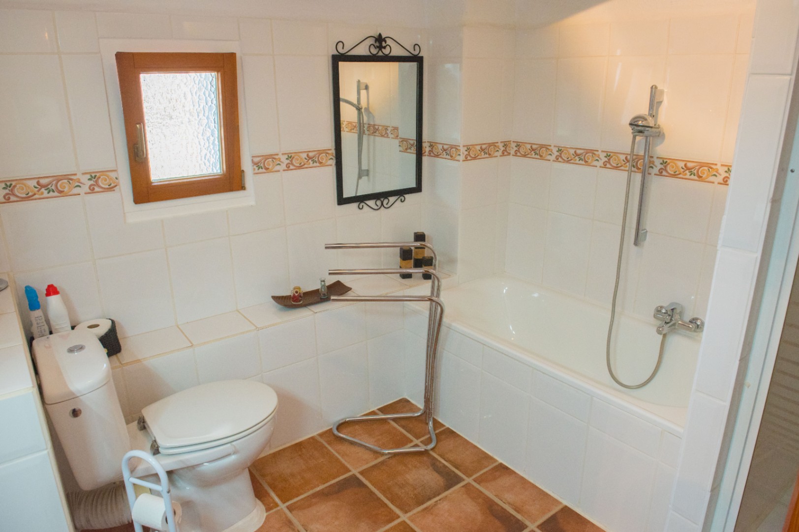 Badkamer met ingebouwd ligbad, terracotta vloertegels en witte wandtegels met een decoratieve rand. Voorzien van een houten raam en smeedijzeren spiegel.