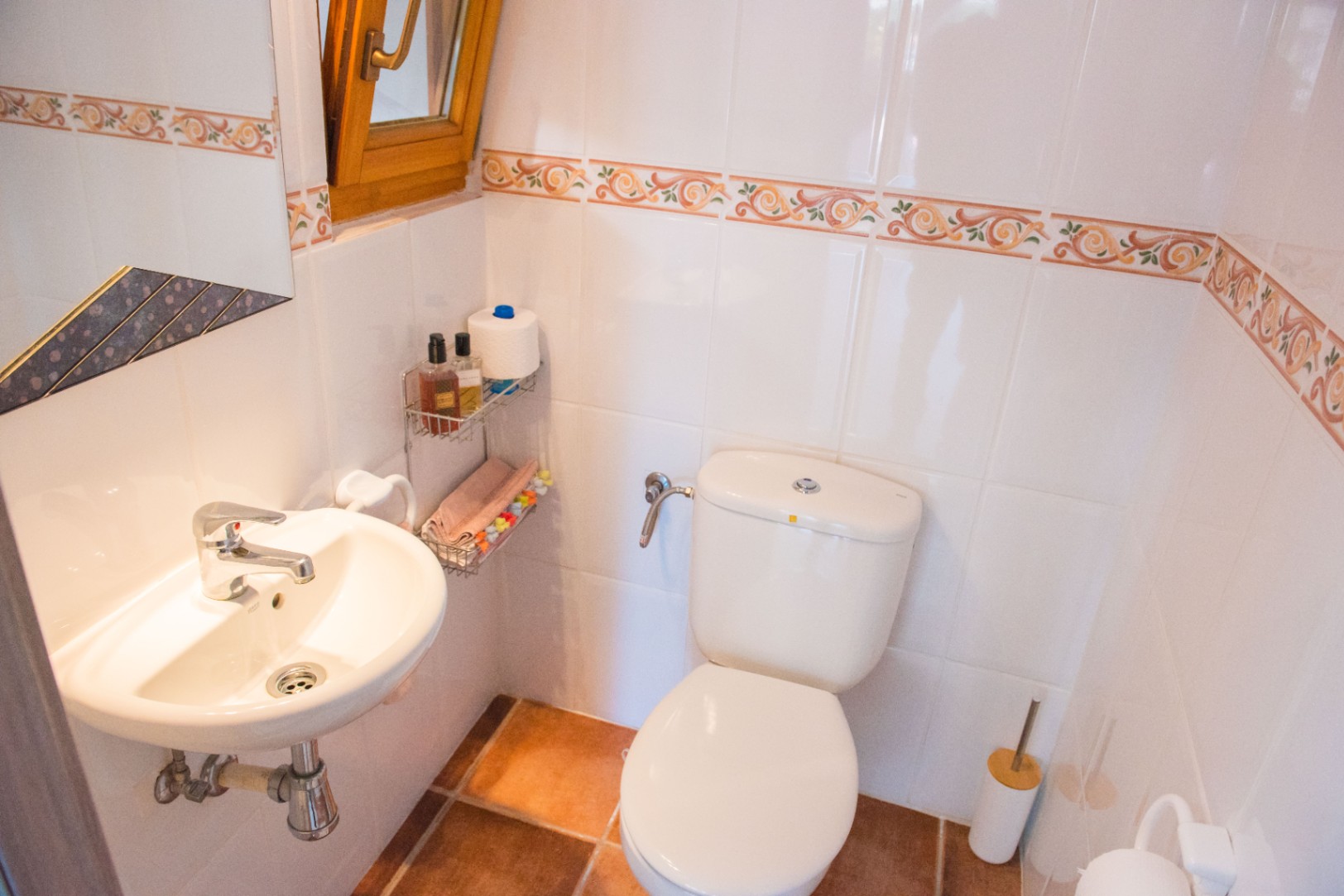 Compacte toiletruimte met witte wandtegels en decoratieve rand, terracotta vloer, wandgemonteerde wastafel en een houten raamkozijn voor natuurlijke ventilatie.