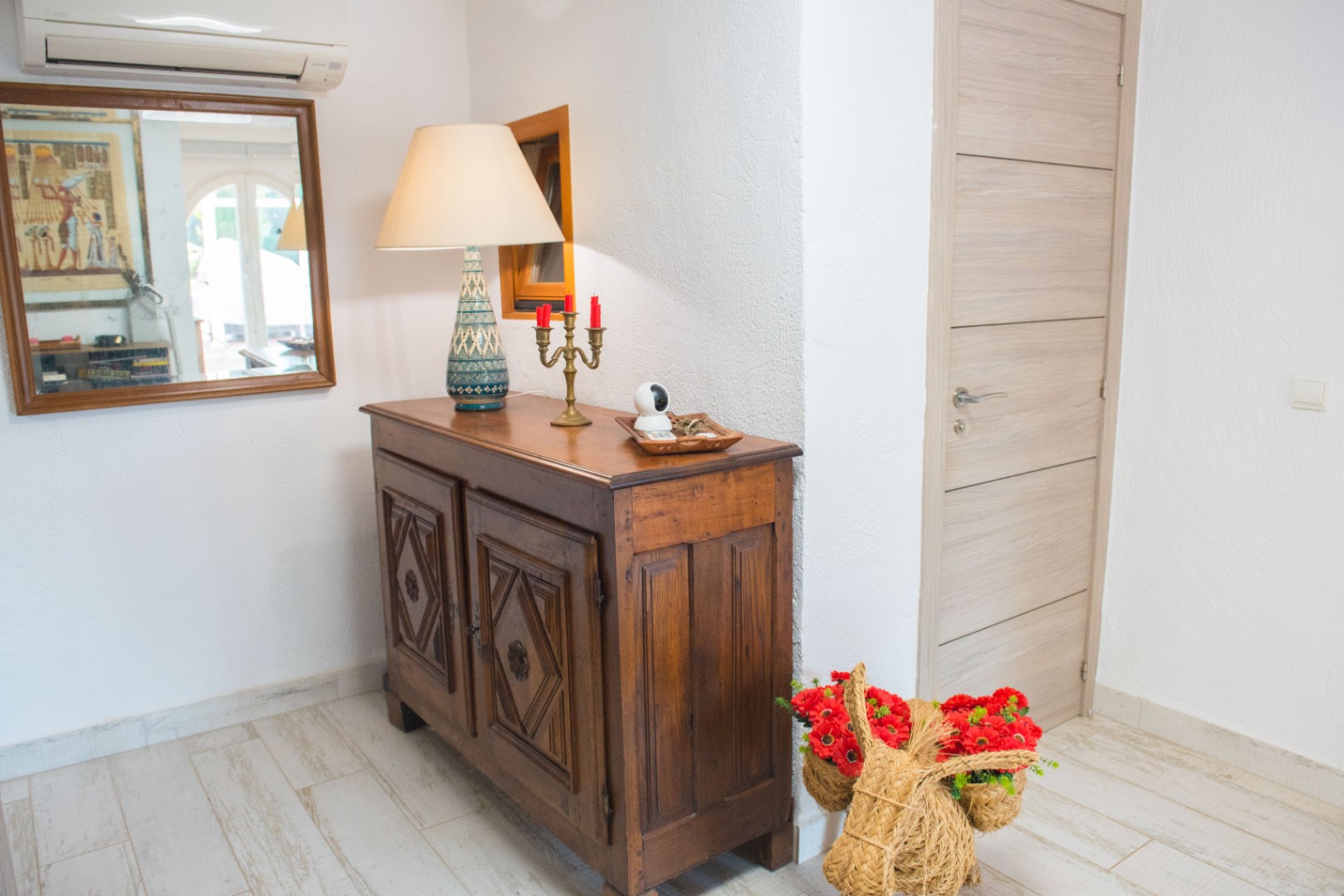 Entree met een bewerkt donkerhouten dressoir, keramische tafellamp en lichte houtlook vloertegels. Voorzien van een spiegel en airconditioning tegen een witte structuurwand.