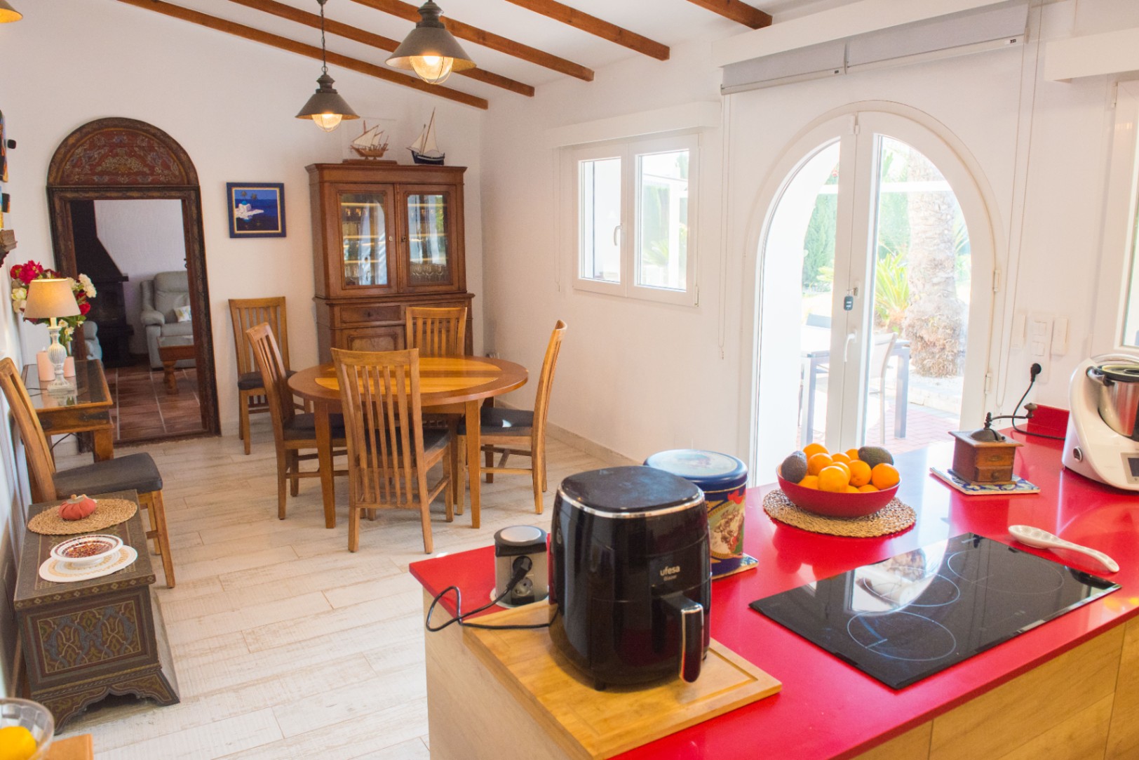 Keuken met rood kookeiland, houten eethoek, zichtbare plafondbalken en een boogvormige glazen deur naar het terras.
