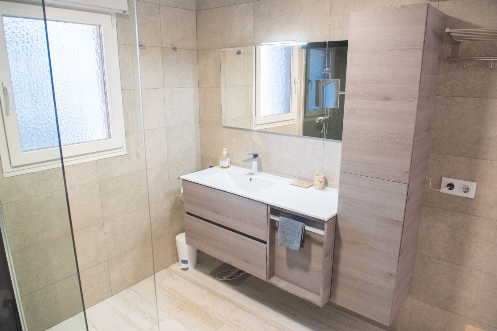 Moderne badkamer met zwevend wastafelmeubel in houtstructuur, bijpassende hoge opbergkast en zandkleurige wandtegels in groot formaat.