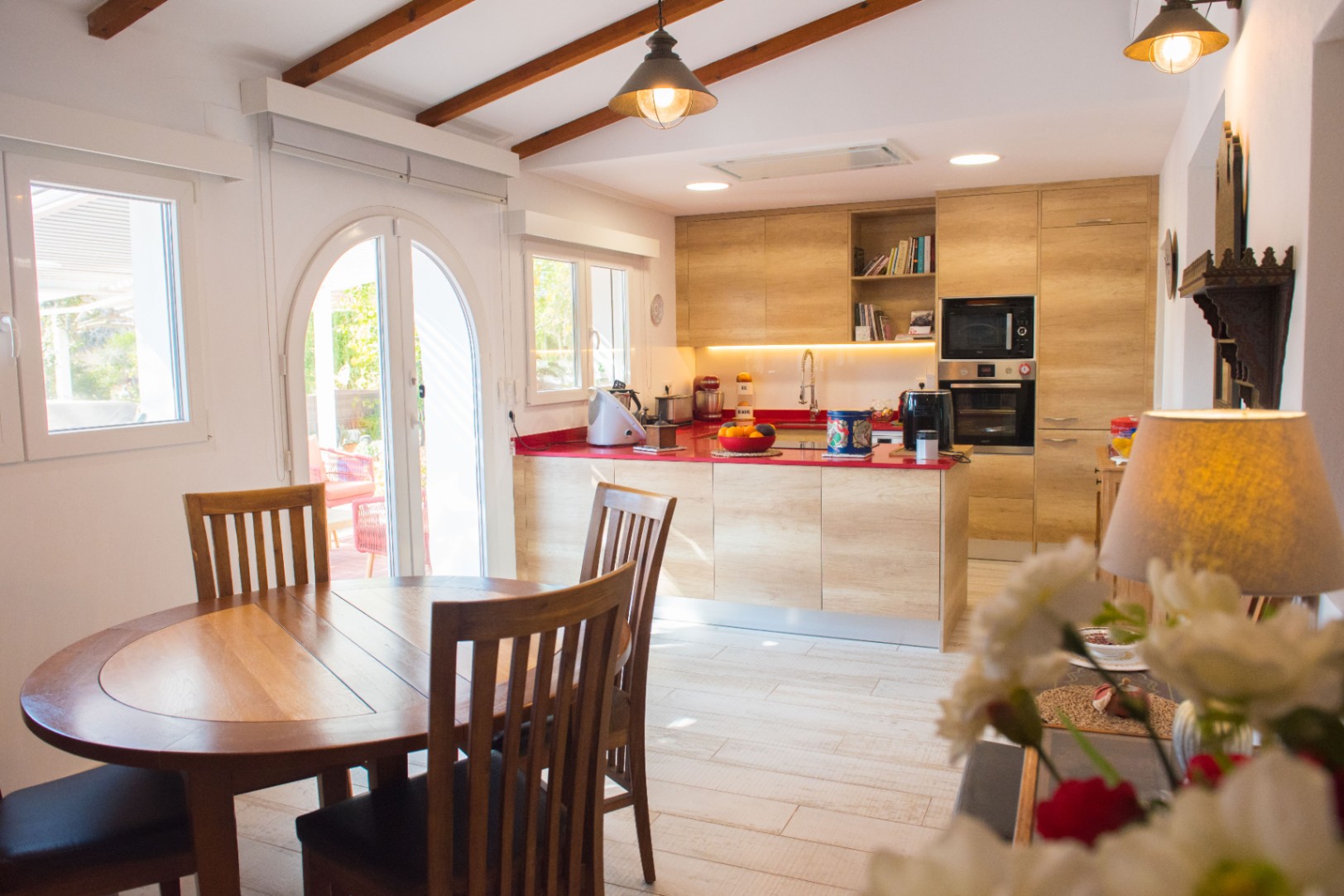 Moderne keuken met lichte houten kasten, een opvallend rood werkblad en een schuin plafond met houten balken, grenzend aan een eethoek met ronde tafel.