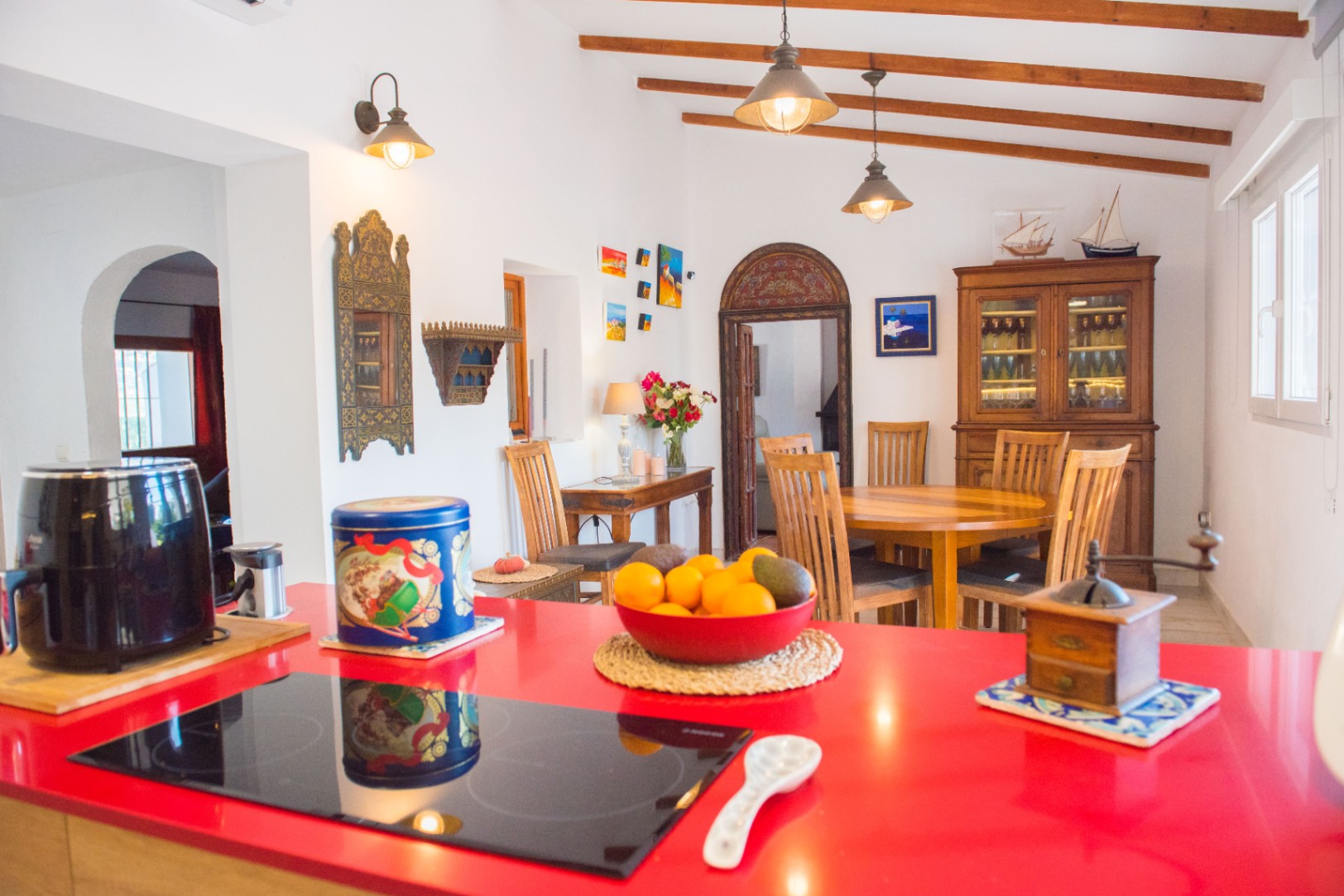 Open keuken met een opvallend rood werkblad en inductiekookplaat, verbonden met een eethoek voorzien van houten plafondbalken en klassiek houten meubilair.