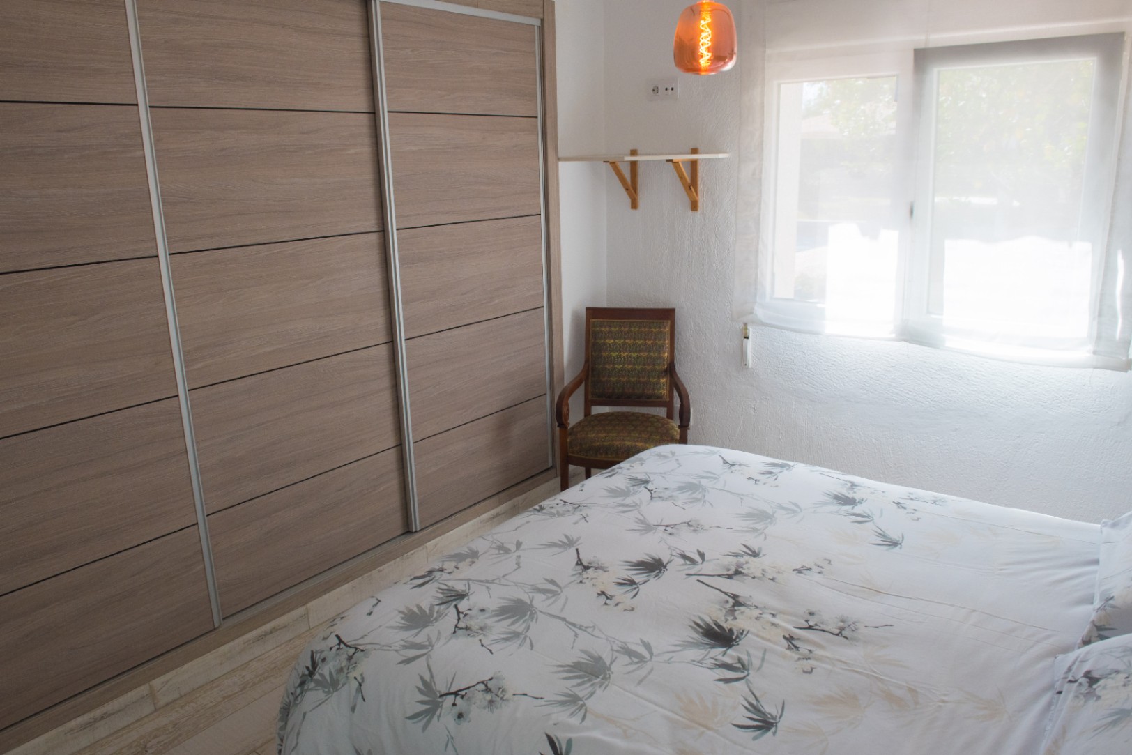 Slaapkamer met een grote inbouwkast met houten schuifdeuren, een vintage fauteuil en natuurlijk lichtinval via het raam met gordijnen.