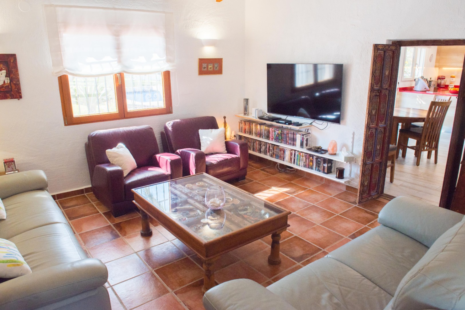 Woonkamer met terracotta tegelvloer, lederen banken en een glazen salontafel. Voorzien van mediaplanken onder de tv en een bewerkte houten deuropening naar de keuken.