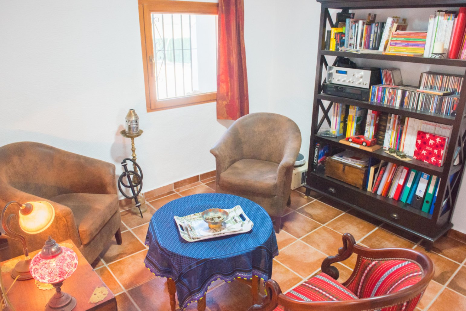 Zithoek met terracotta tegelvloer, een donkerhouten boekenkast en fauteuils rond een ronde tafel met een blauw tafelkleed.