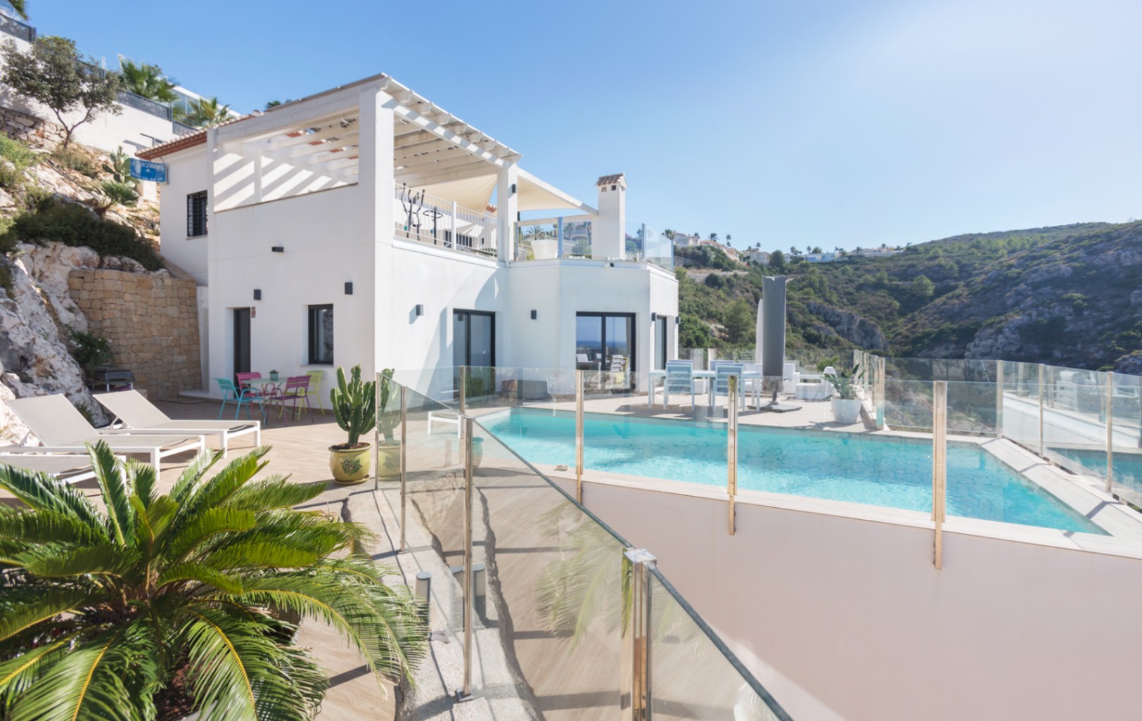 Buitenaanzicht van een witte villa met meerdere niveaus, een zwembad met glazen omheining, zonneterrassen en panoramisch uitzicht op de heuvels.