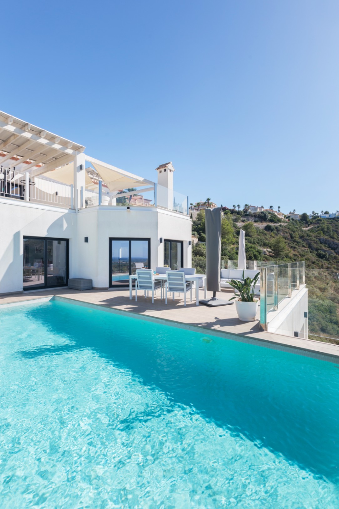 Infinity zwembad met turquoise water grenzend aan een betegeld terras met eethoek en glazen balustrades met uitzicht op de heuvels.