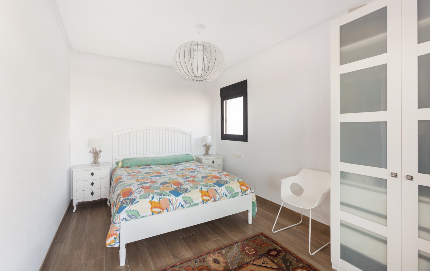Lichte slaapkamer met wit houten bedframe, bijpassende nachtkastjes en een kledingkast met matglas. Voorzien van een houtlook vloer en een moderne design hanglamp.